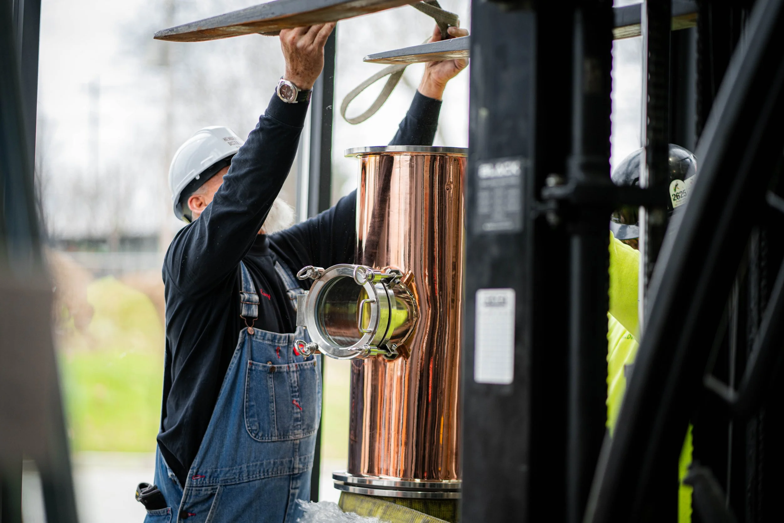 A worker wearing a white safety helmet, black shirt, and denim overalls is installing or inspecting a large, shiny copper pipe using a lift system. The worker's face is not visible.