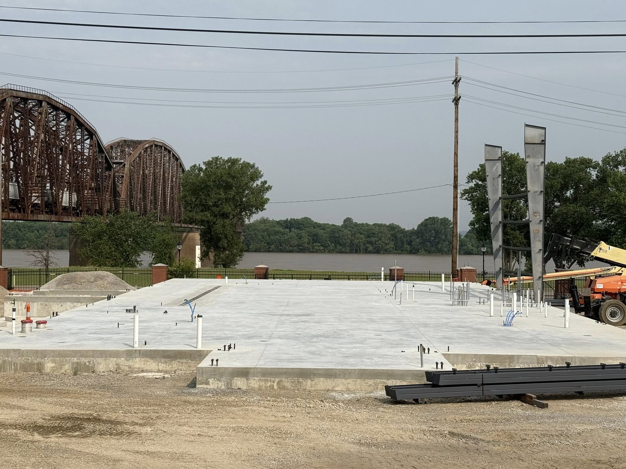 Construction site with concrete foundation, pipes, and structural elements under development near a river, with a bridge and trees in the background.