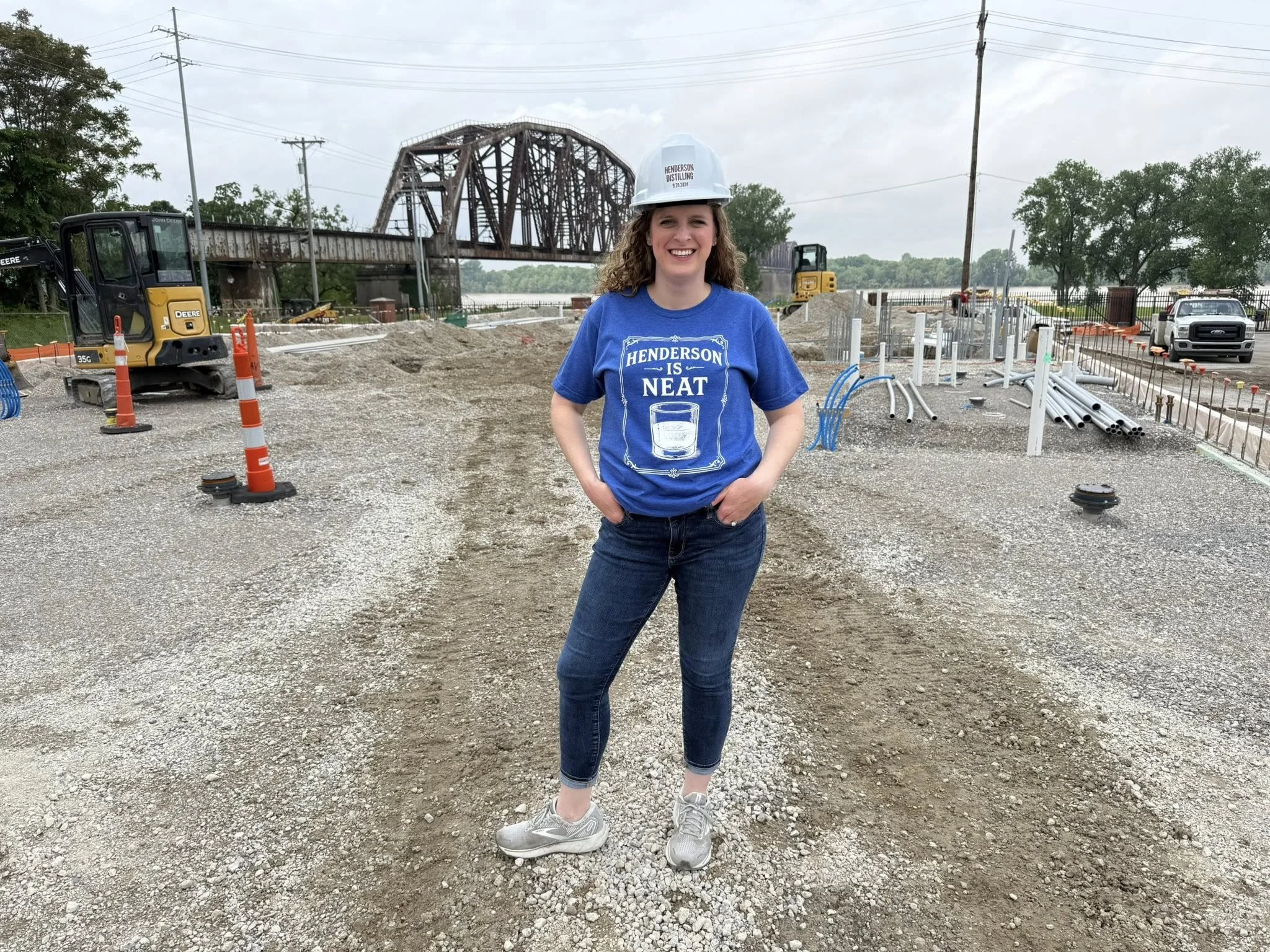 A woman wearing a white construction helmet and a blue t-shirt with a glass of Henderson distilling logo, standing on a construction site near a bridge and construction equipment.