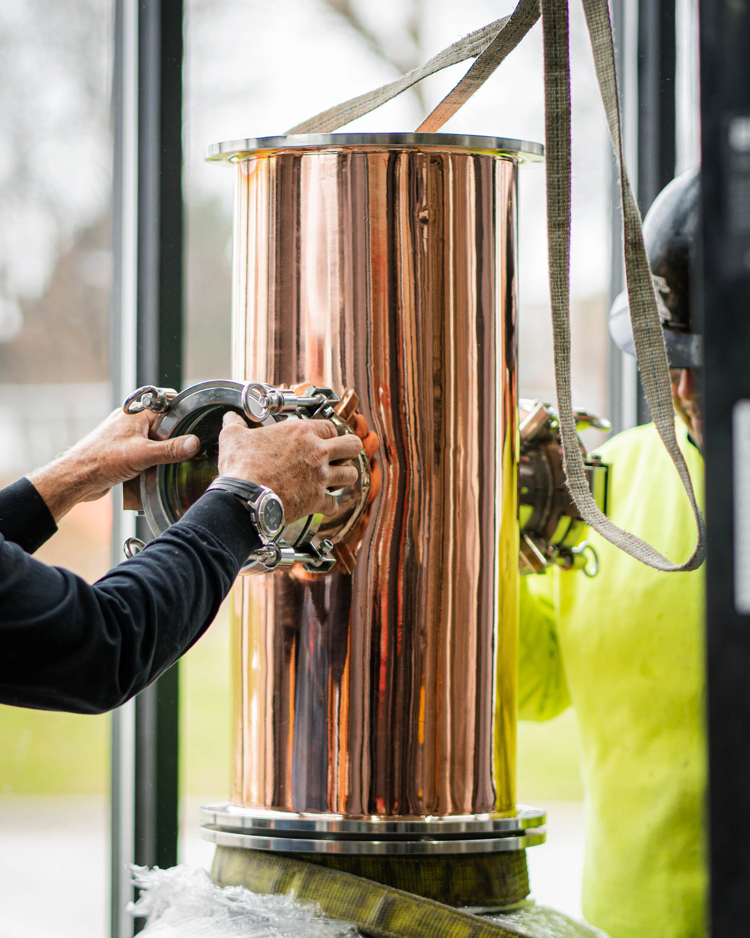 Close-up of a person handling a large, shiny, copper-colored cylindrical vessel with metallic fittings, in a bright environment, likely a laboratory or industrial setting.