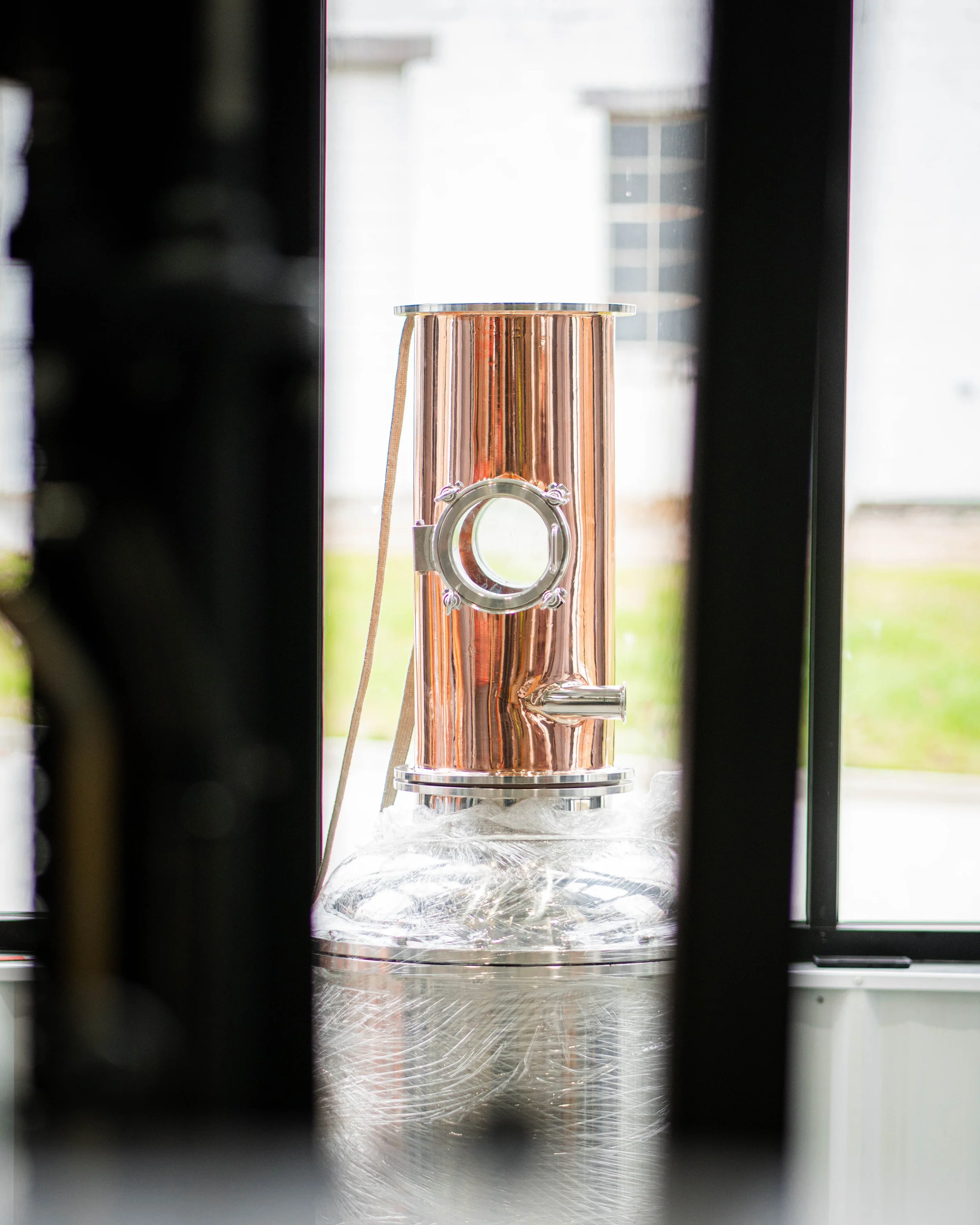 A copper still on a table seen through a window with black framing.