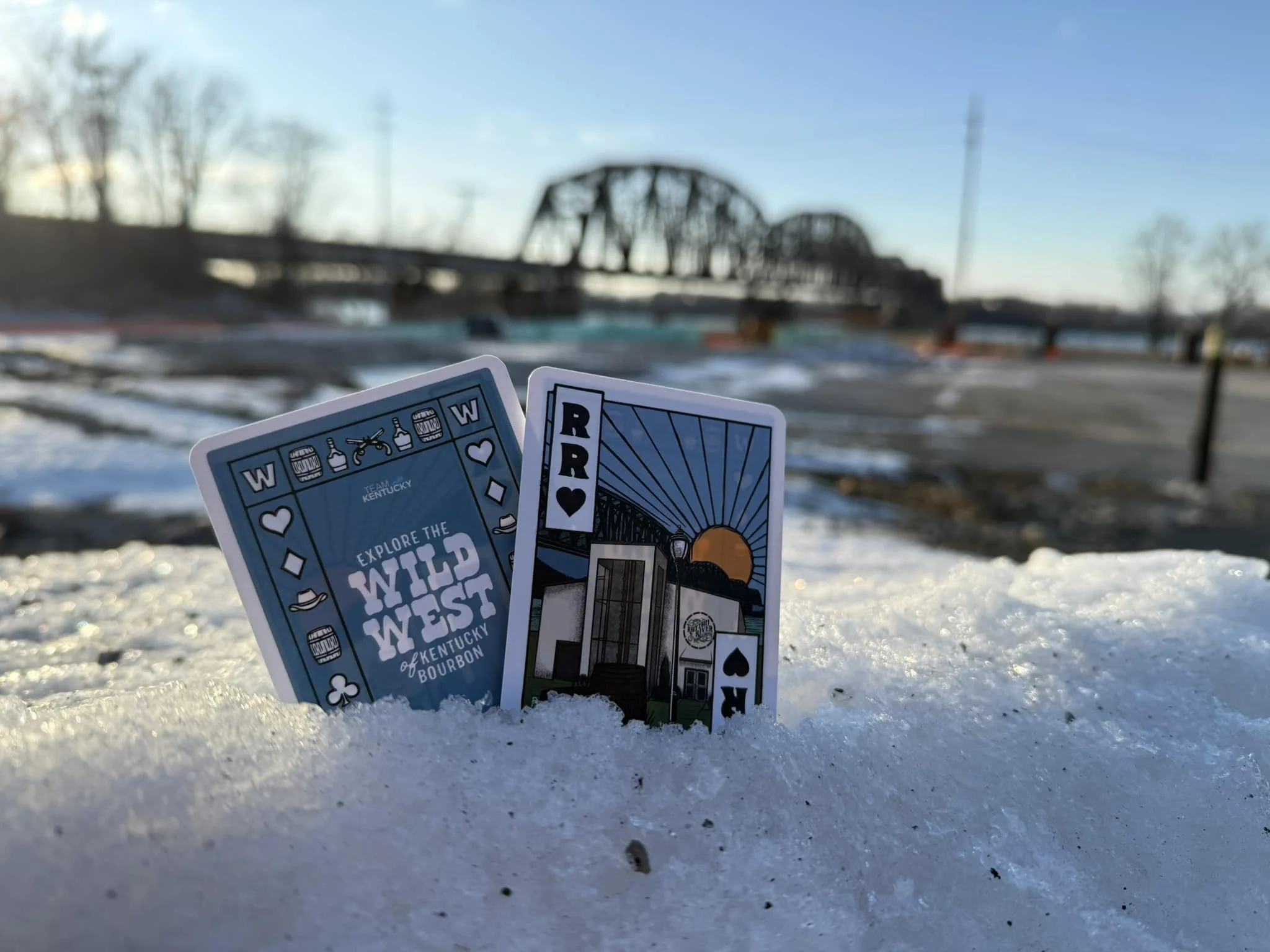 Two playing cards stuck in snow with a bridge in the background during sunset or sunrise.