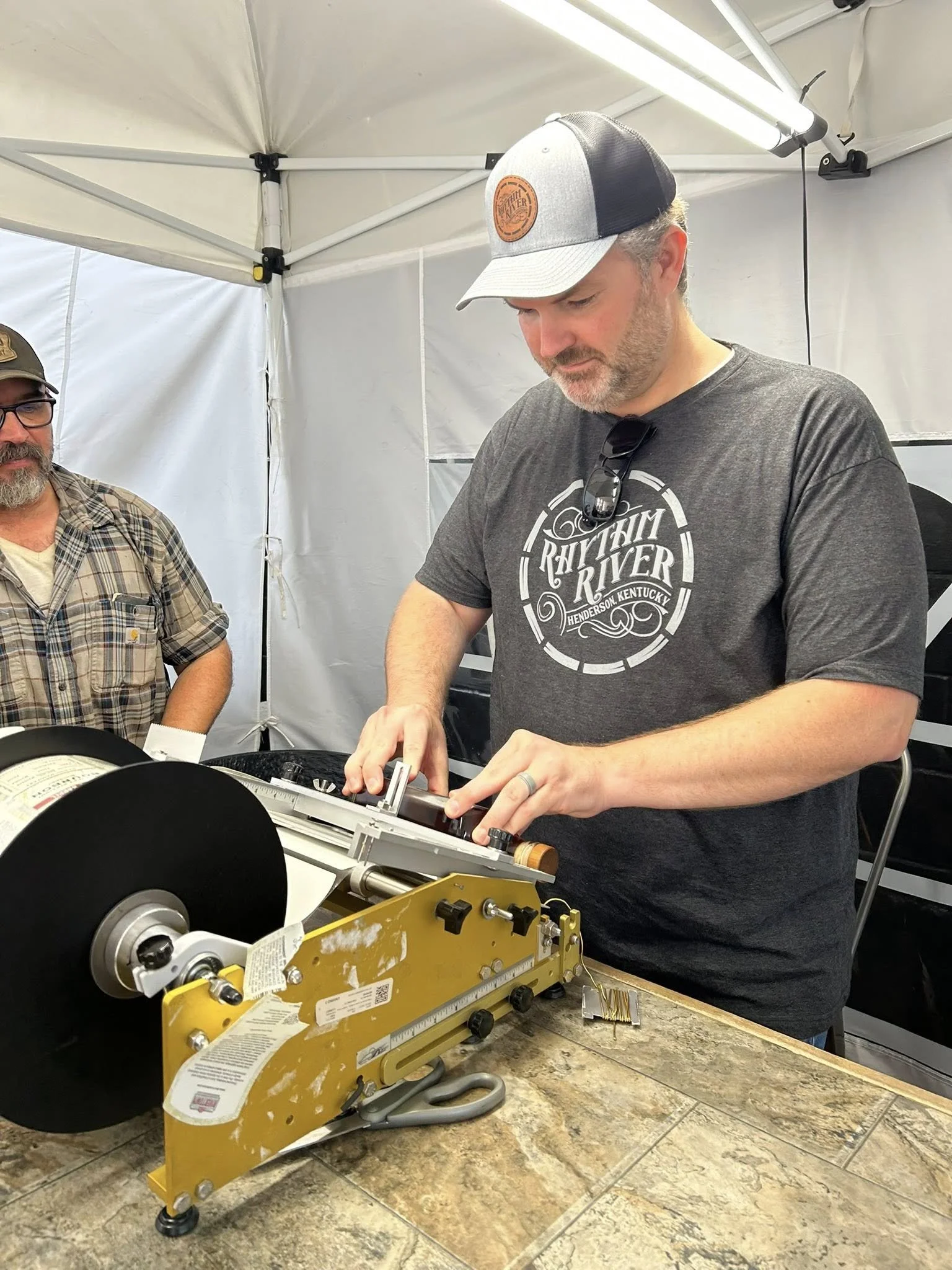 A man wearing a gray and black baseball cap and a dark gray T-shirt, with sunglasses hanging from his shirt, is operating a paper cutter or guillotine, while another man in a plaid shirt and glasses looks on inside a white tent