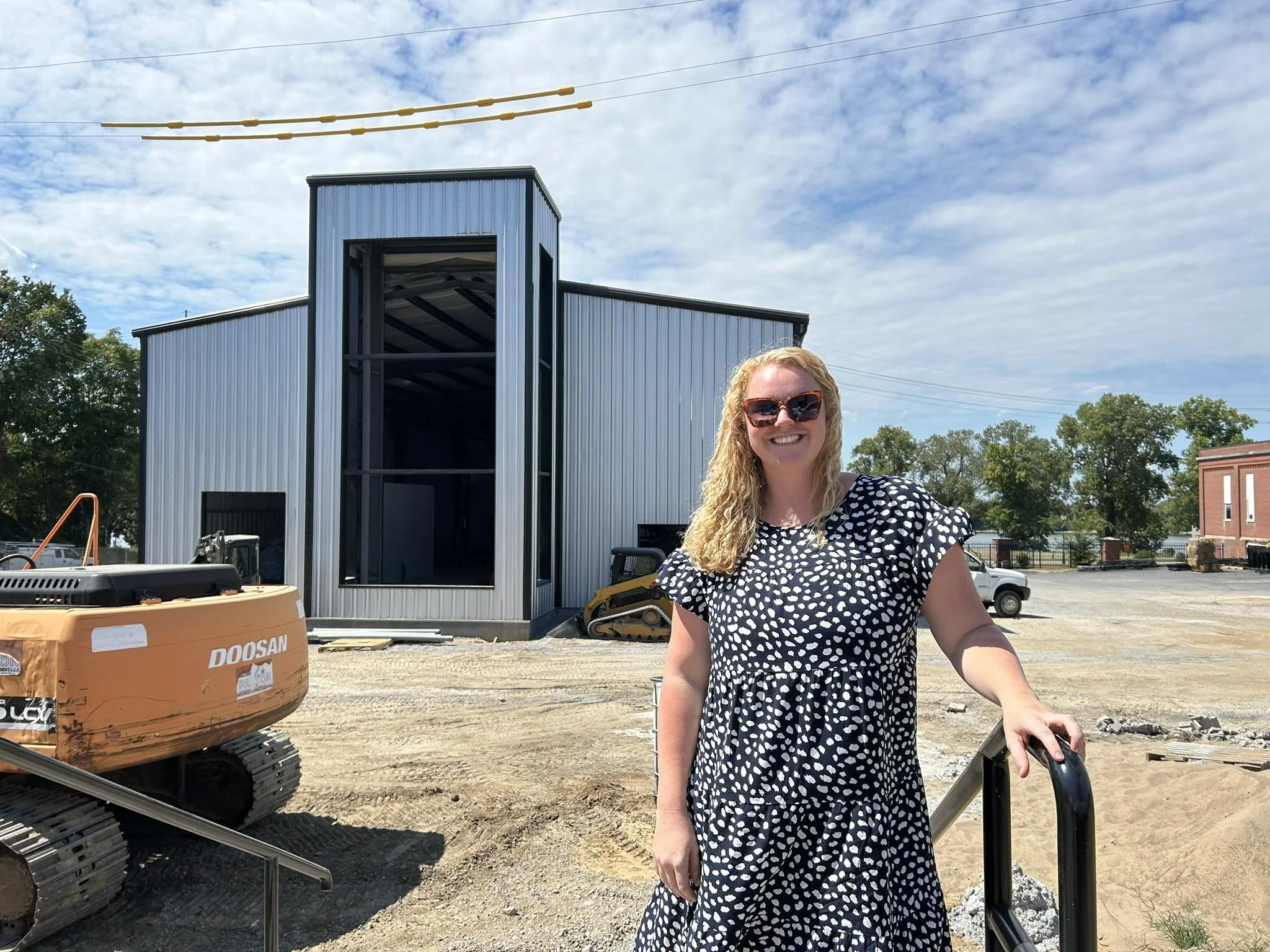 A woman with blonde curly hair wearing sunglasses and a black and white polka dot dress, smiling while standing with her hand on a railing in front of a construction site. There is a large metal building under construction, a construction excavator, 