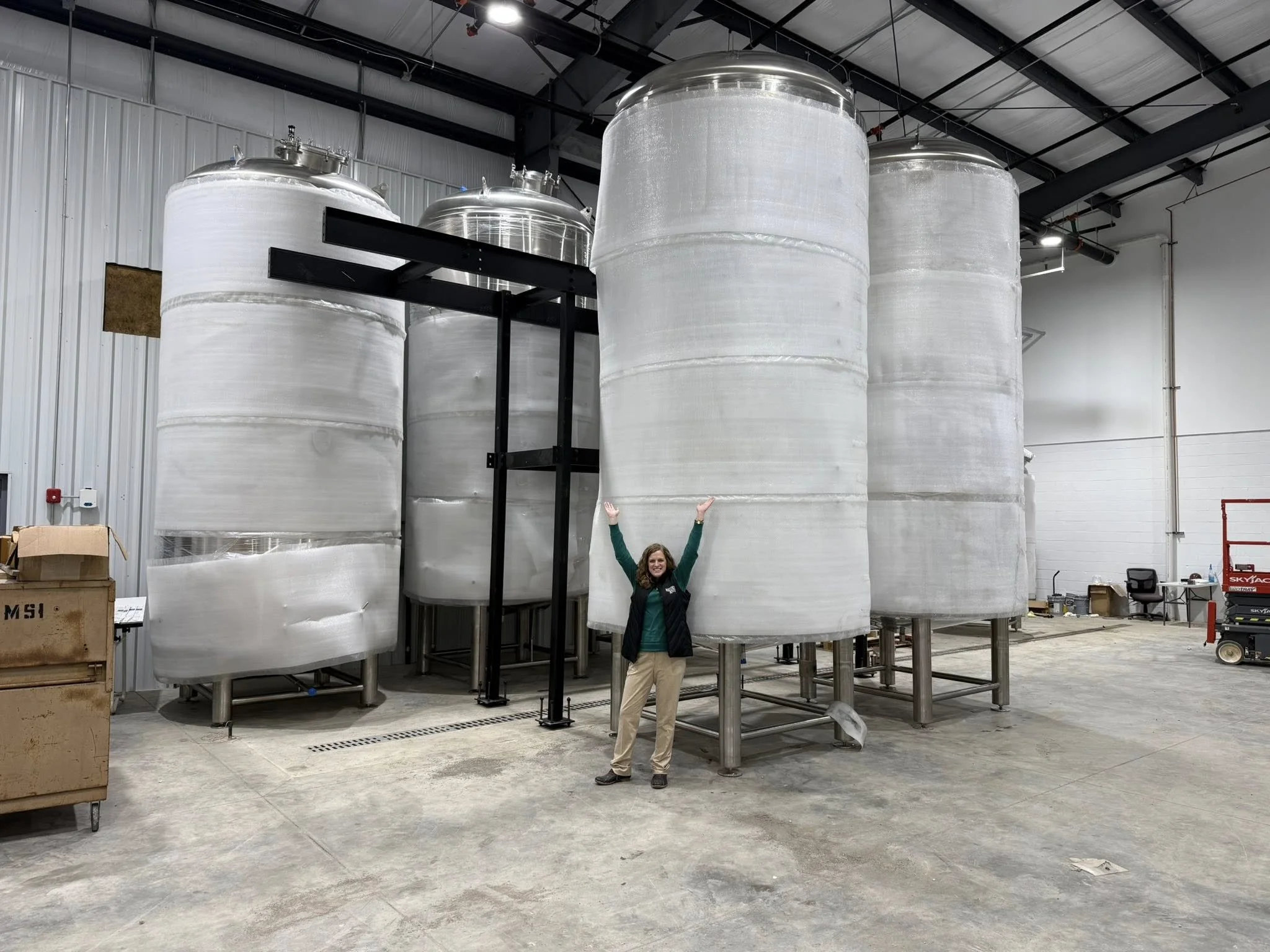 A woman standing in front of large metal storage tanks inside a warehouse, raising her arms in celebration.