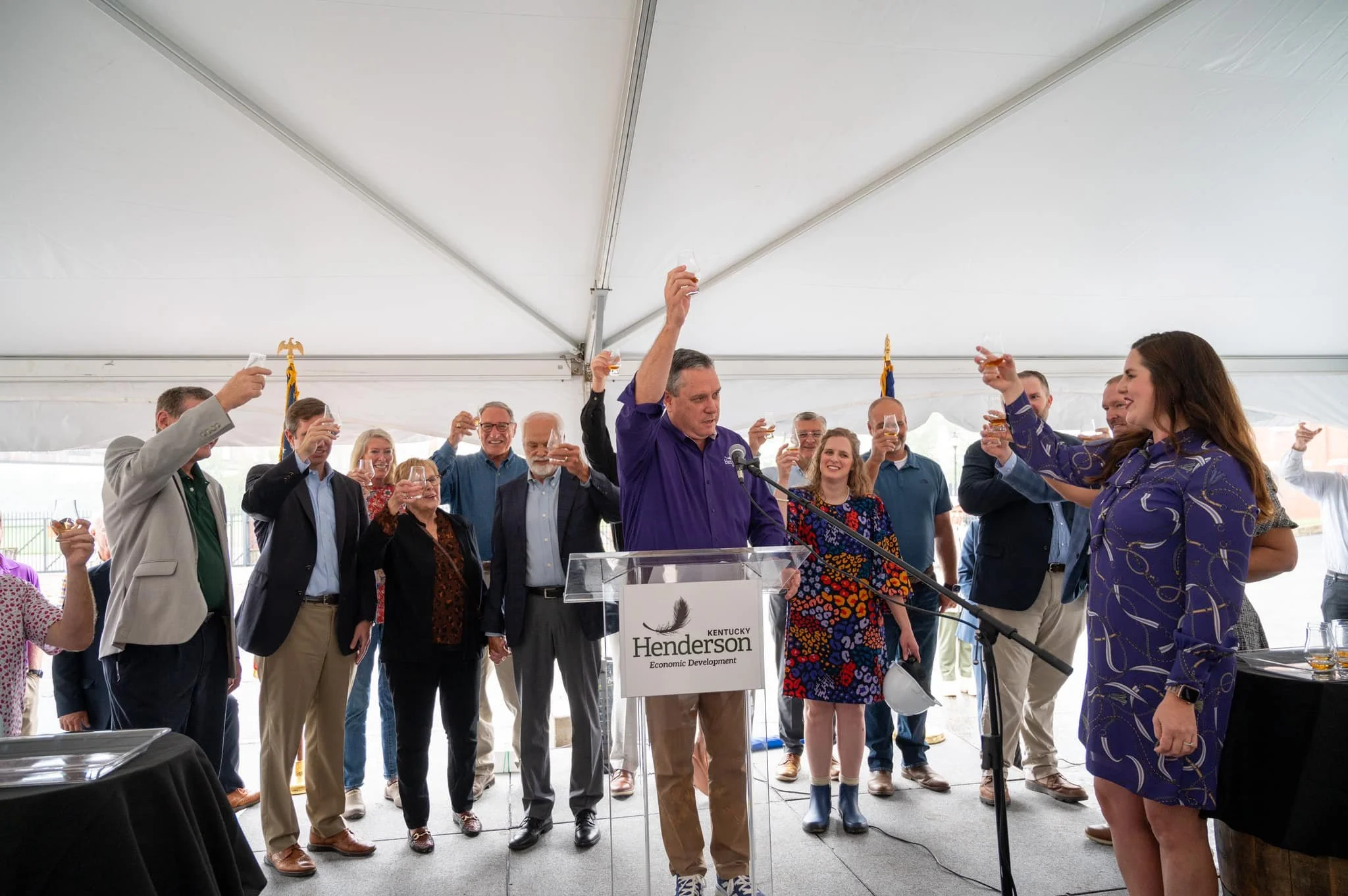Group of people celebrating under a tent, holding glasses for a toast, with a man at a clear podium labeled 'Kentucky Henderson Economic Development'.