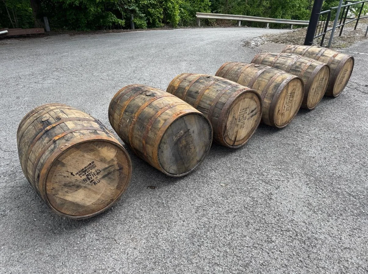 Six wooden barrels lying on their sides on a paved surface near a grassy, wooded area with a metal railing.