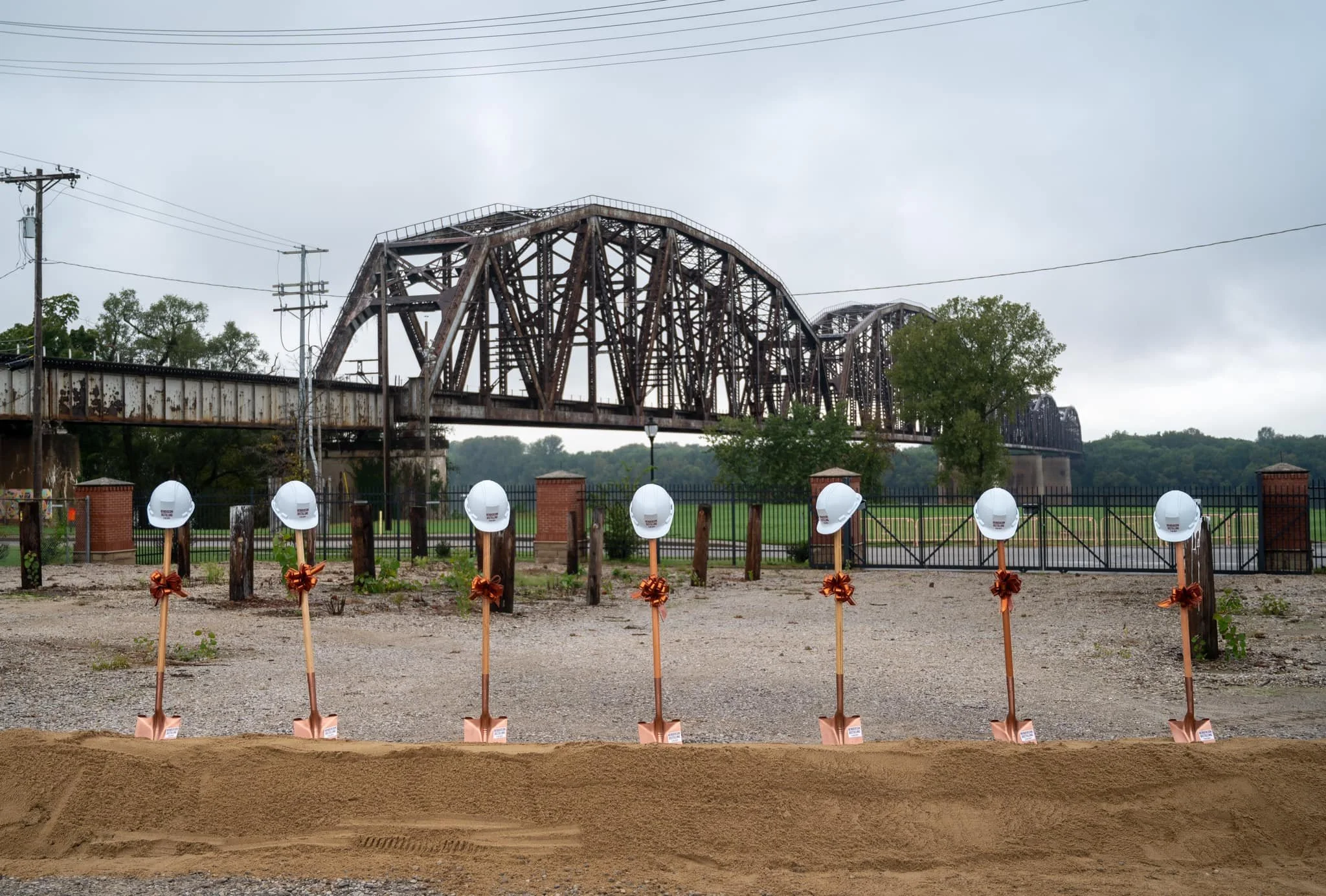 A groundbreaking ceremony with seven shovels topped with white safety helmets, decorated with orange ribbons, in front of a historic rusted train bridge under cloudy skies, with a fence and green trees in the background.