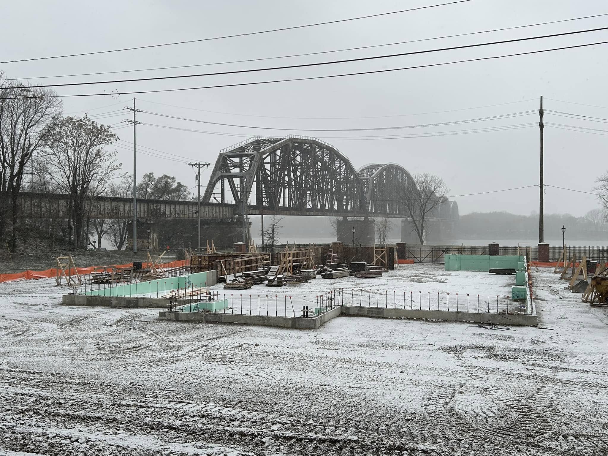 Construction site with a foundation and rebar, snow on the ground, in front of a bridge over a river on a foggy day with leafless trees and utility poles.