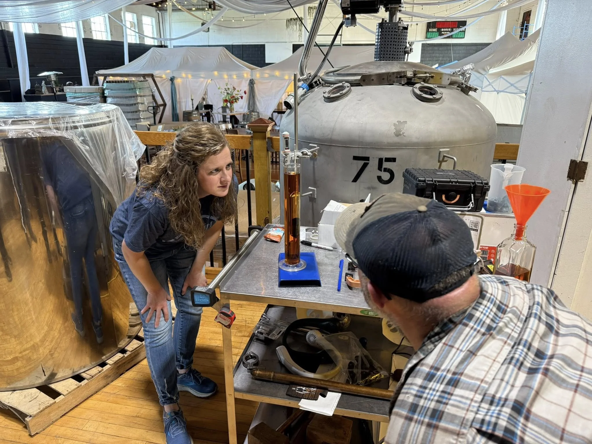 A woman and a man observing a tall graduated cylinder filled with a dark liquid on a table, in a distillery or brewery setting, with equipment and storage tanks in the background.