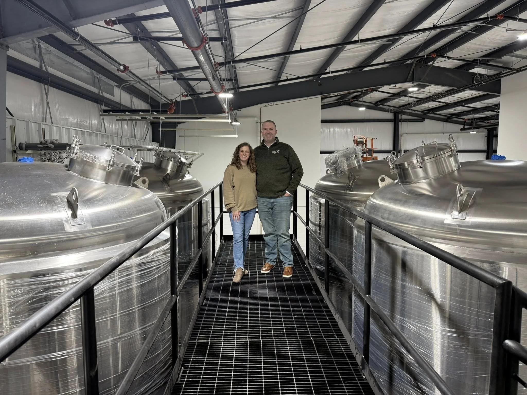 Two smiling people, a woman and a man, standing on a metal walkway inside a brewery or industrial facility, surrounded by large stainless steel tanks.