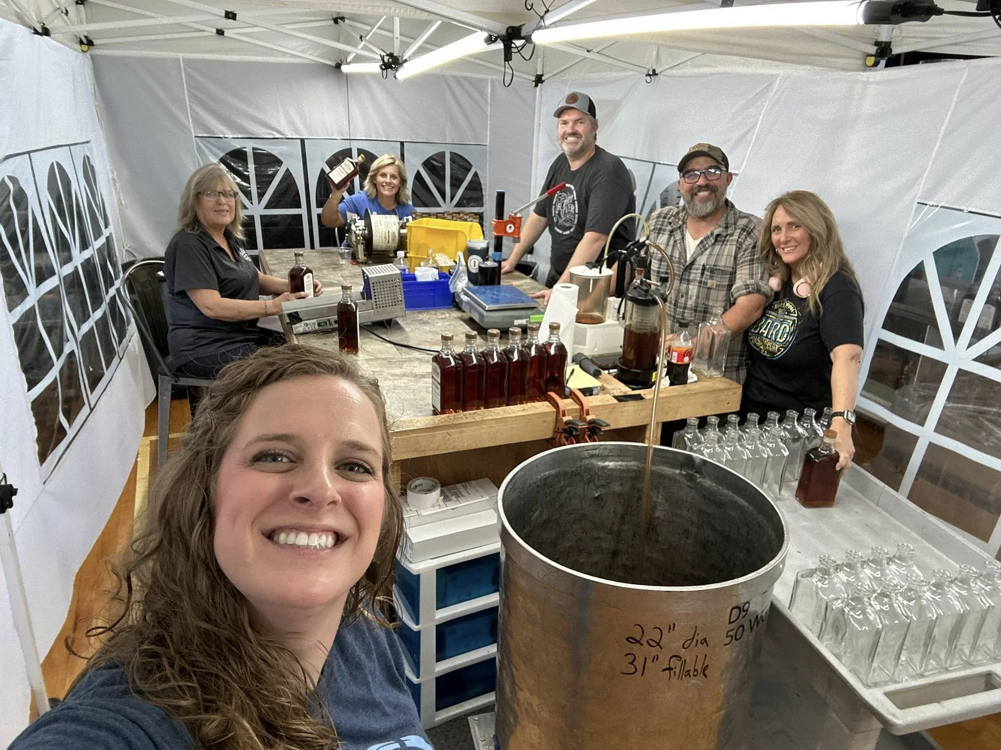 Group of people inside a white tent making bottles of liquor, with a smiling woman taking a selfie in the foreground.