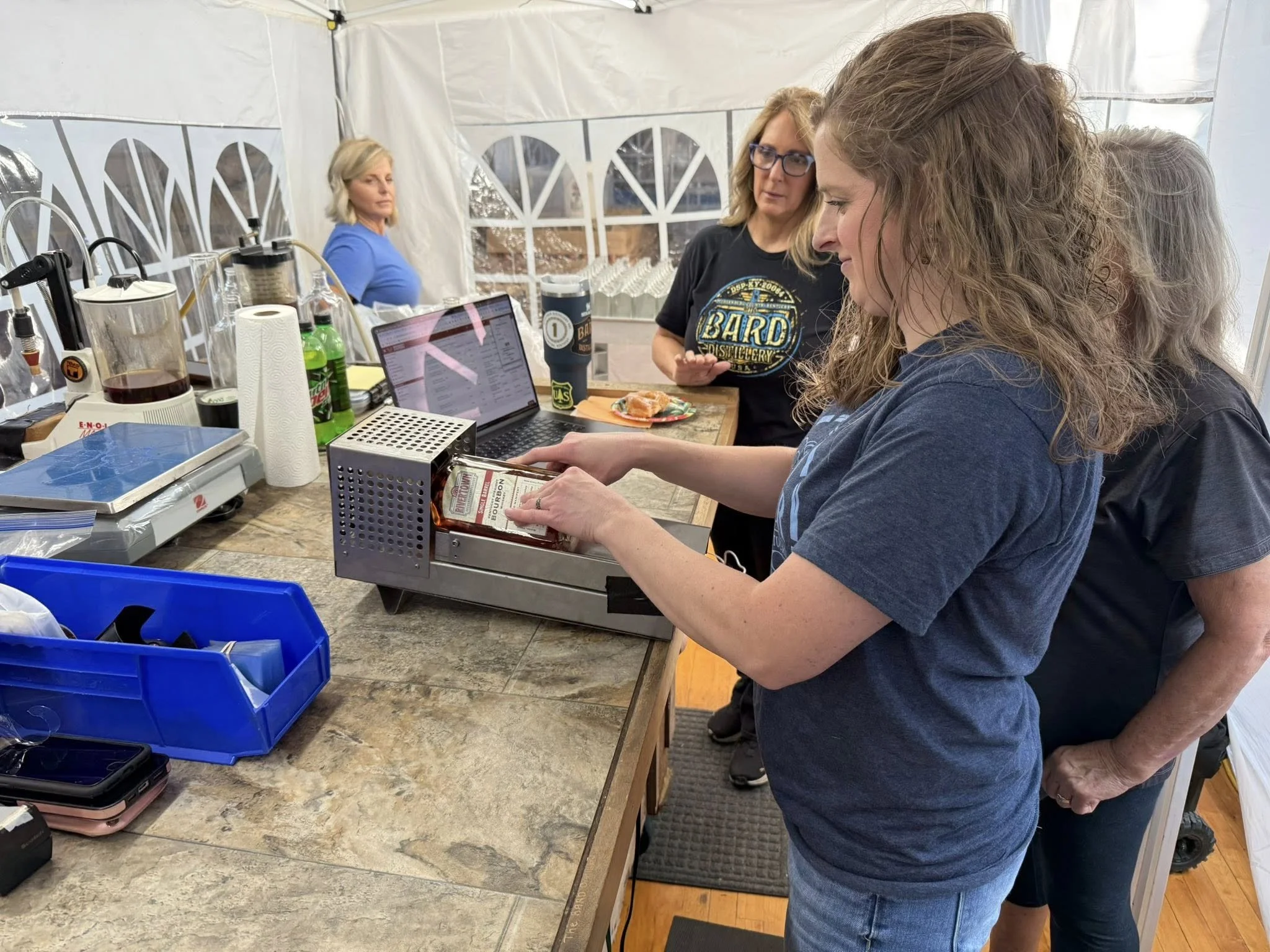 Women in a brewery or distillery working at a countertop with equipment and a laptop, one woman inserting a bottle of whiskey into a bottle filler.