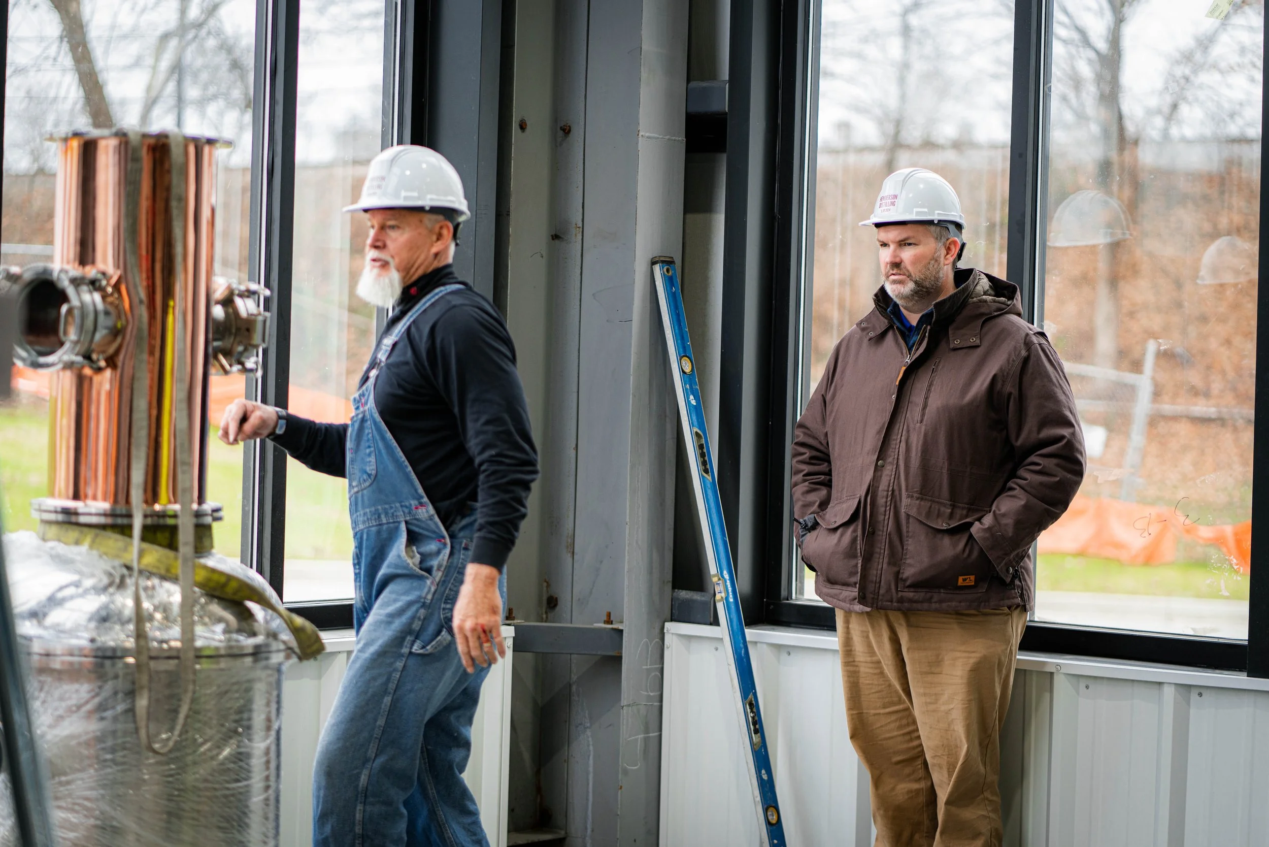 Two men wearing safety helmets inside a building with large windows; one is inspecting machinery, the other stands observing, with a ladder leaning against the wall.