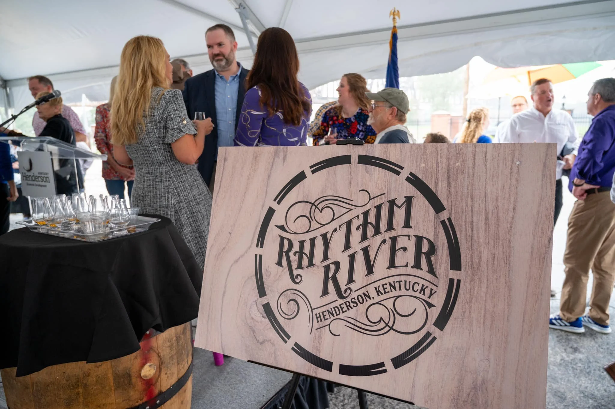 People gathered at an event under a large tent, with a sign reading 'Rhythm River Henderson, Kentucky' in the foreground, and a table with tasting glasses nearby.