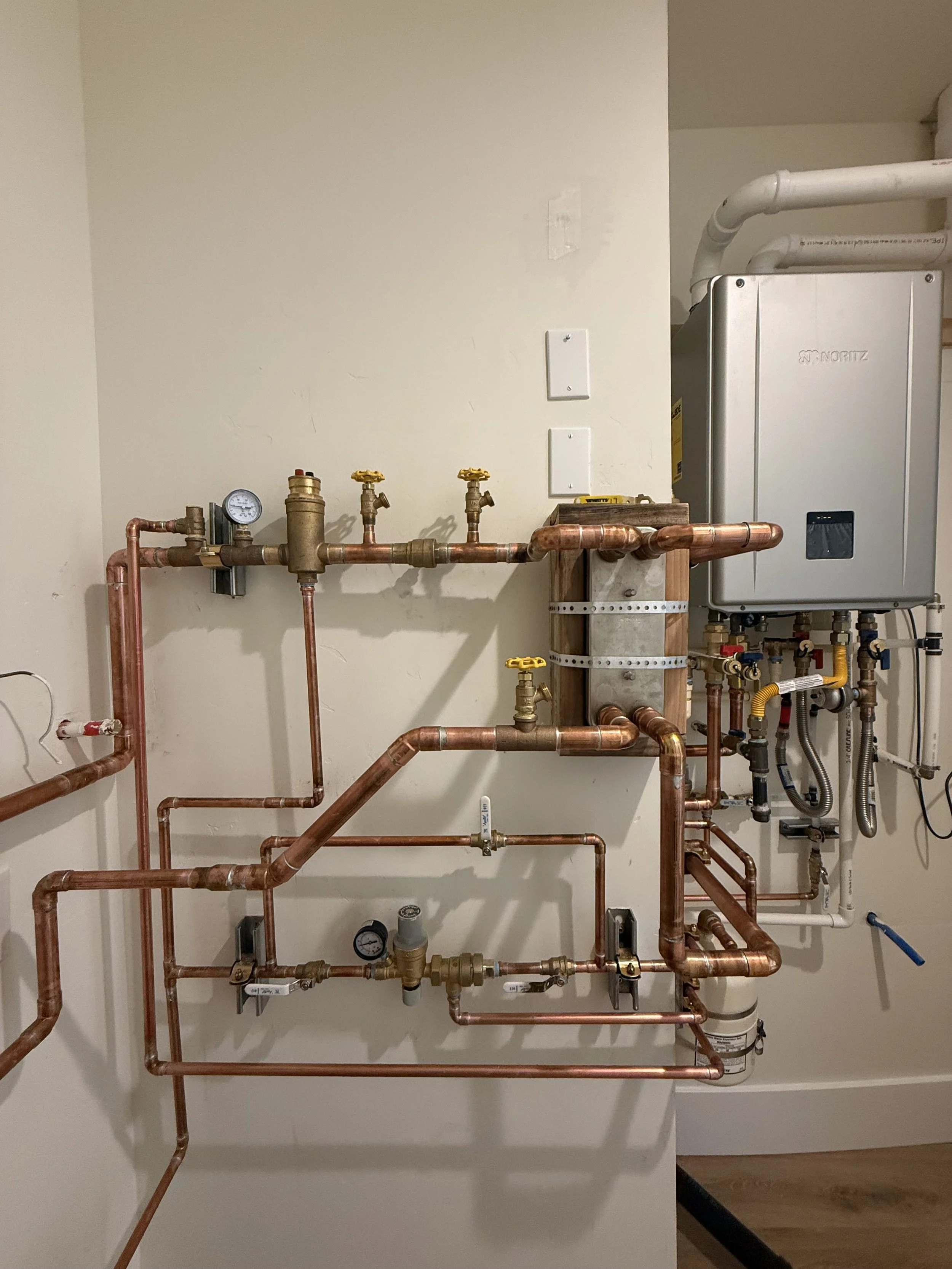 Complex plumbing system with copper pipes, gauges, valves, and a tankless water heater mounted on the wall.