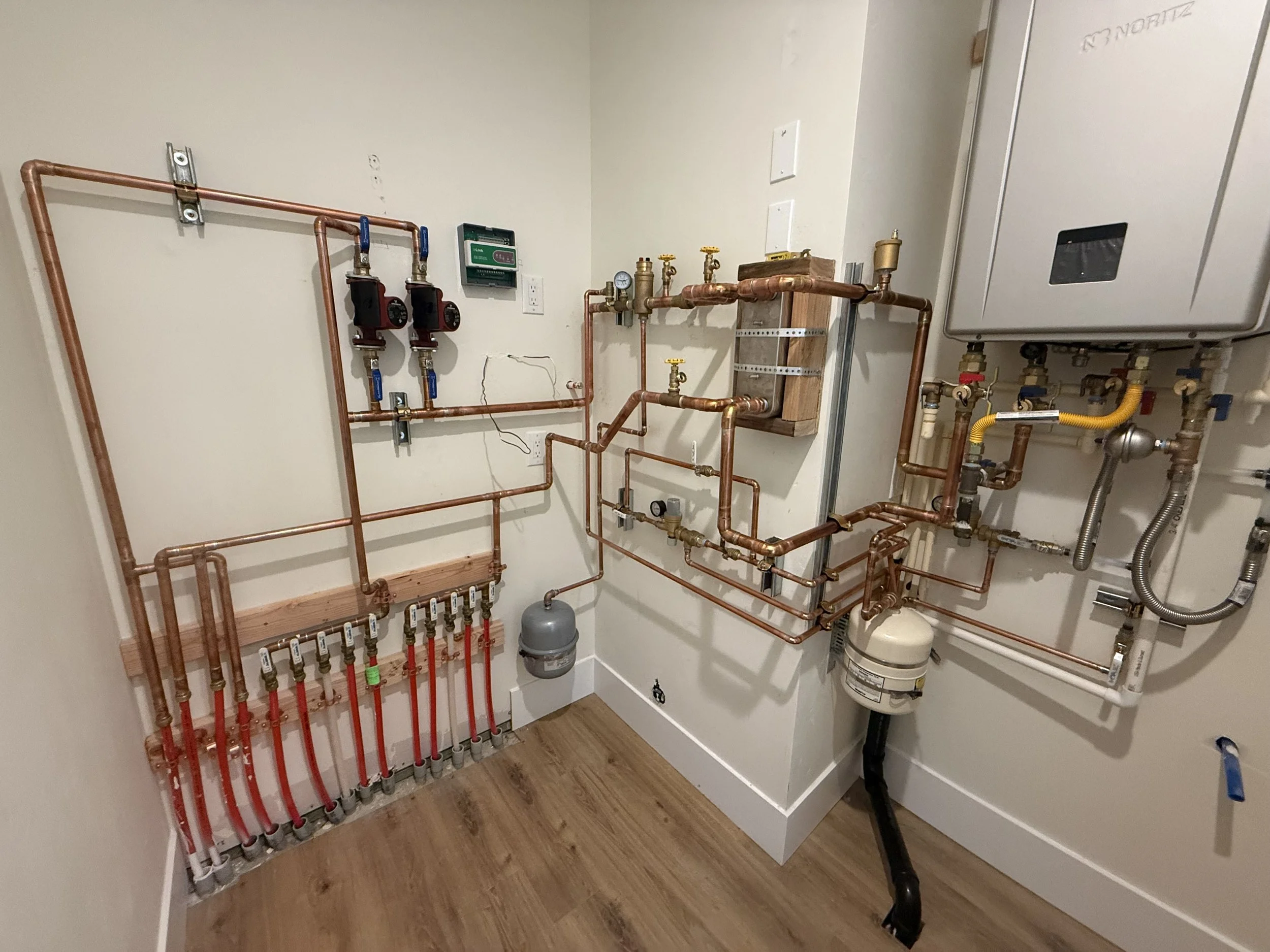 A plumbing room with copper pipes, valves, gauges, and a boiler system, installed along the wall with floor-mounted piping and flooring.