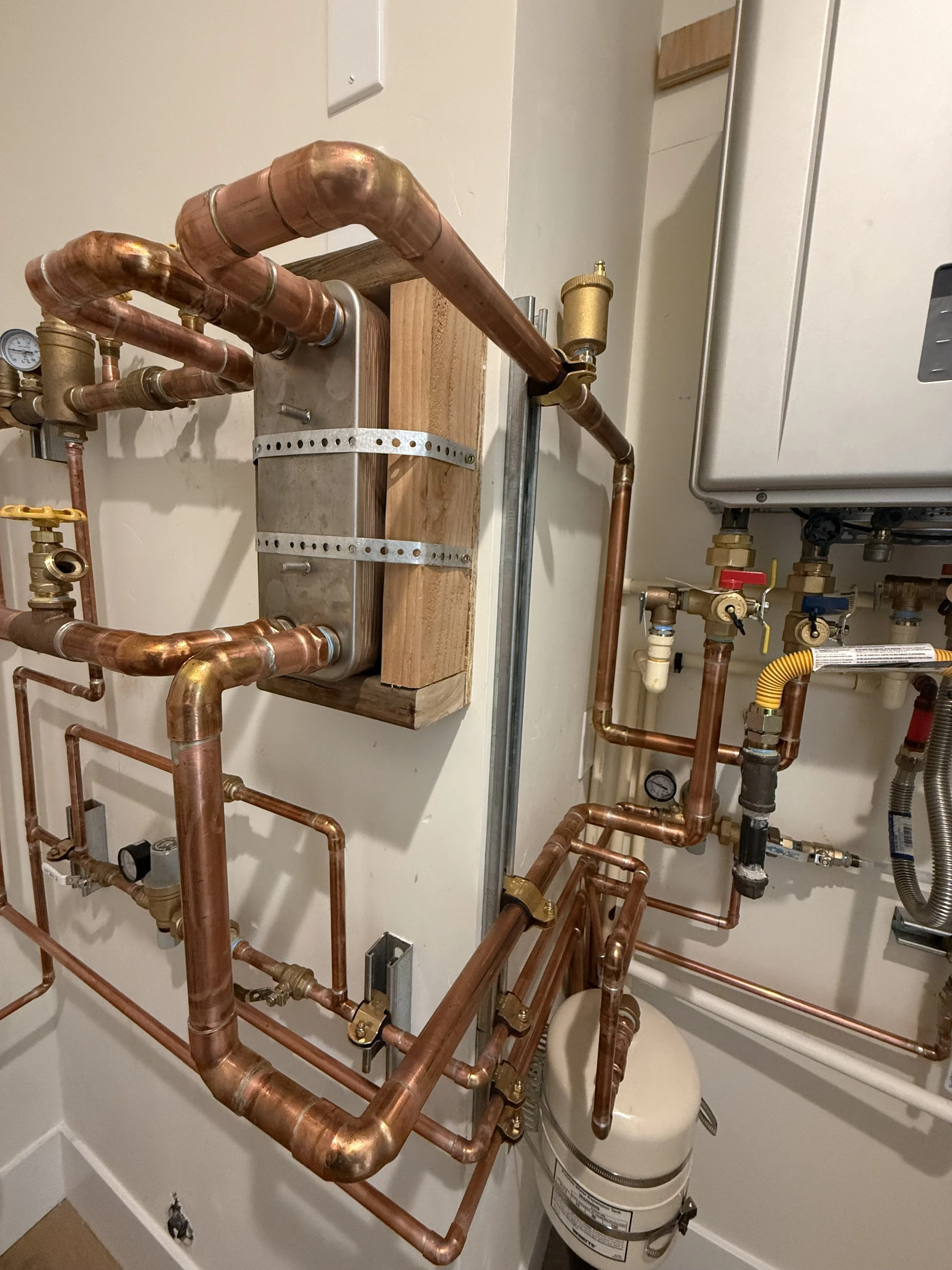 A plumbing system with copper pipes and fittings, some pipes connected to a large tank, mounted on a wall, with various valves and gauges.