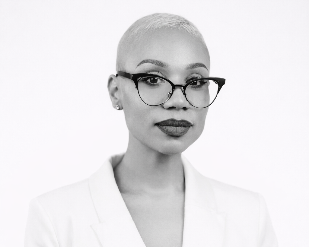 Black and white portrait of a woman with short platinum blonde hair, glasses, lipstick, and earrings, wearing a white blazer, against a plain light background.