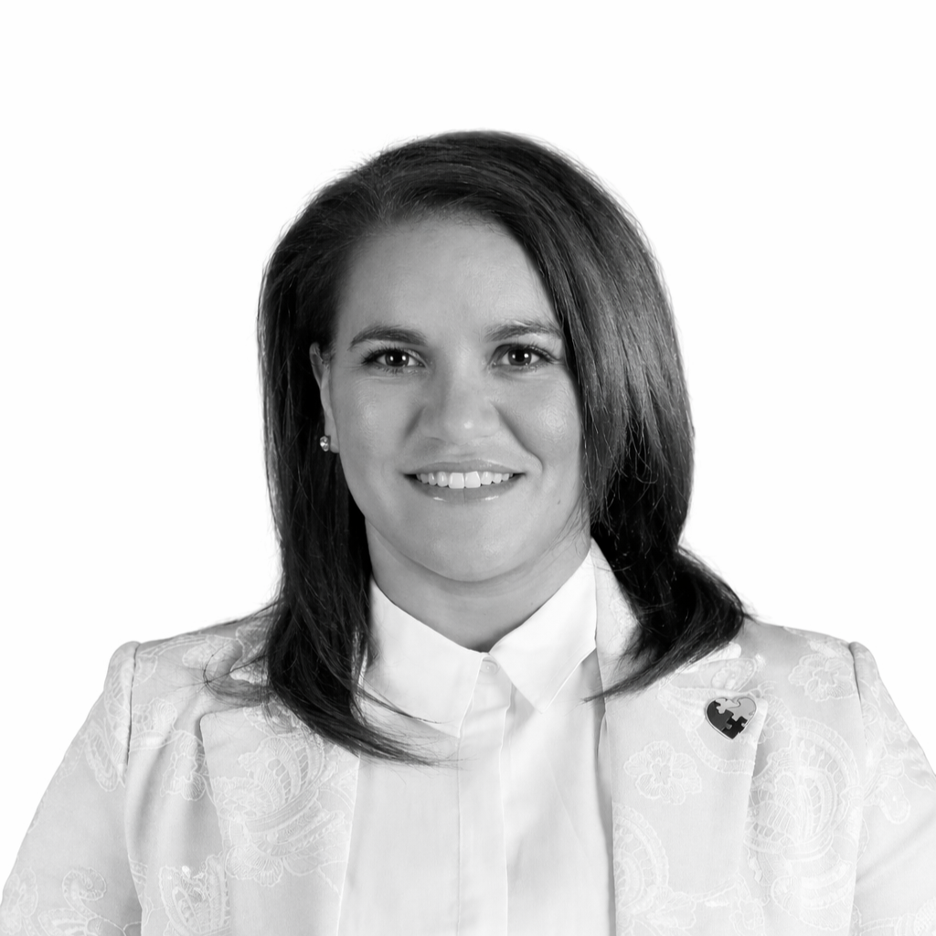Black and white portrait of a smiling woman with shoulder-length dark hair, wearing a white blouse and a floral-patterned jacket with a puzzle piece heart pin.