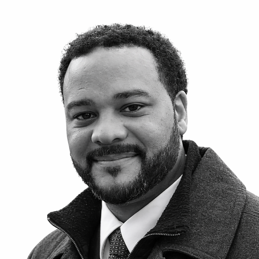 Close-up of a smiling man with curly hair and beard, wearing a suit and tie.