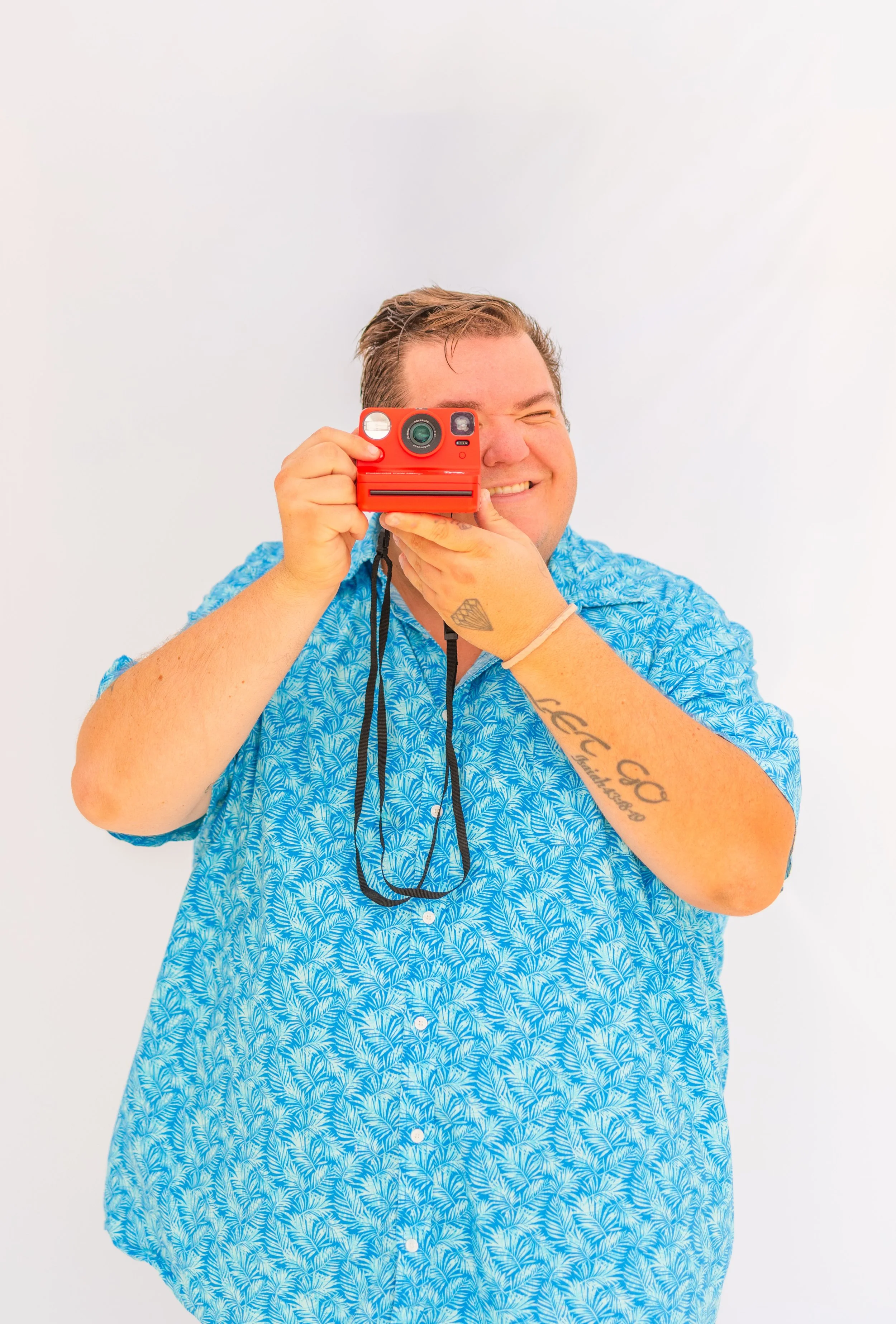A man in a blue patterned shirt smiling and holding a red camera, taking a selfie against a white background.