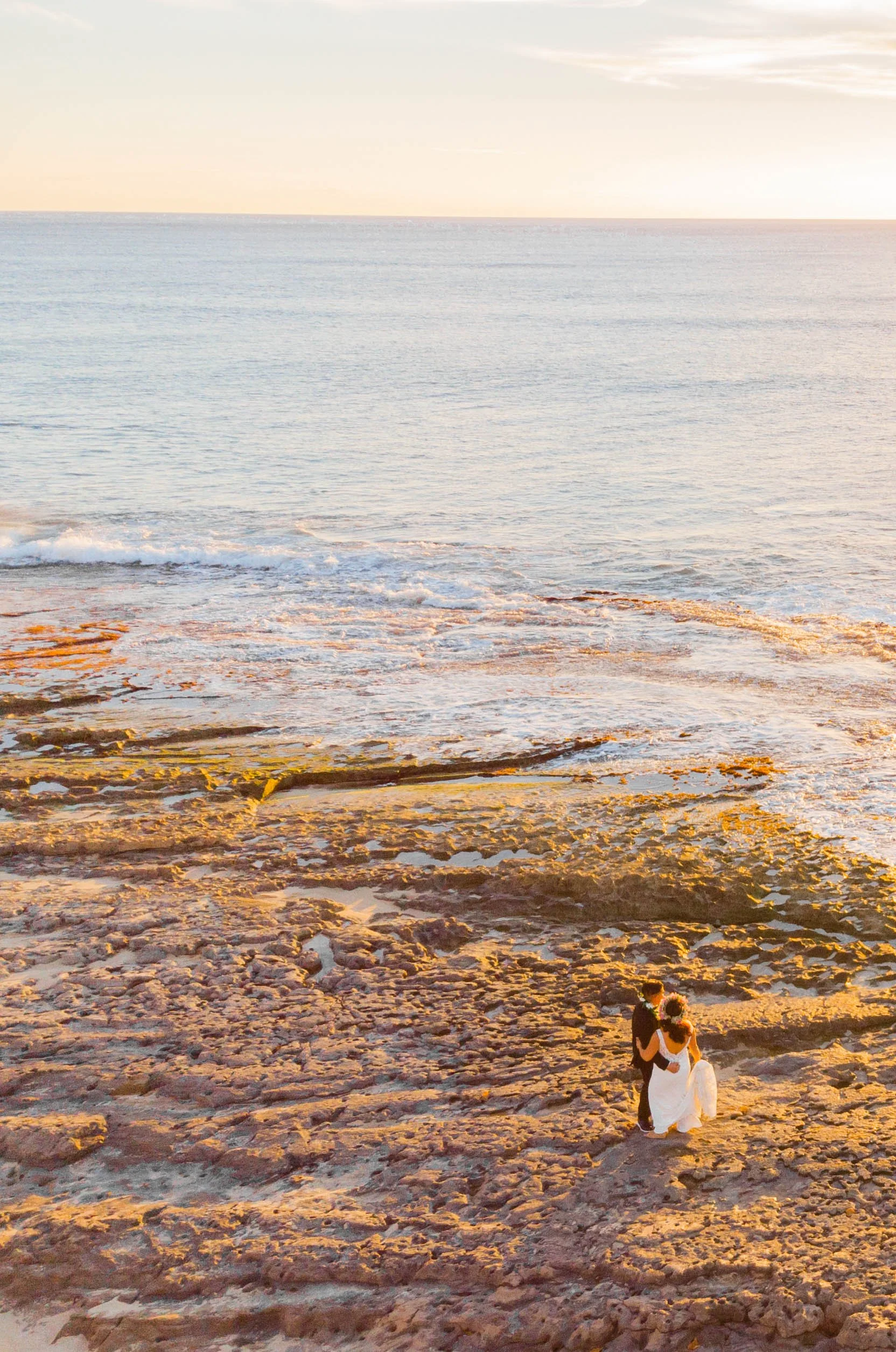 A bride and groom standing on rocky beach at sunset, facing the ocean.