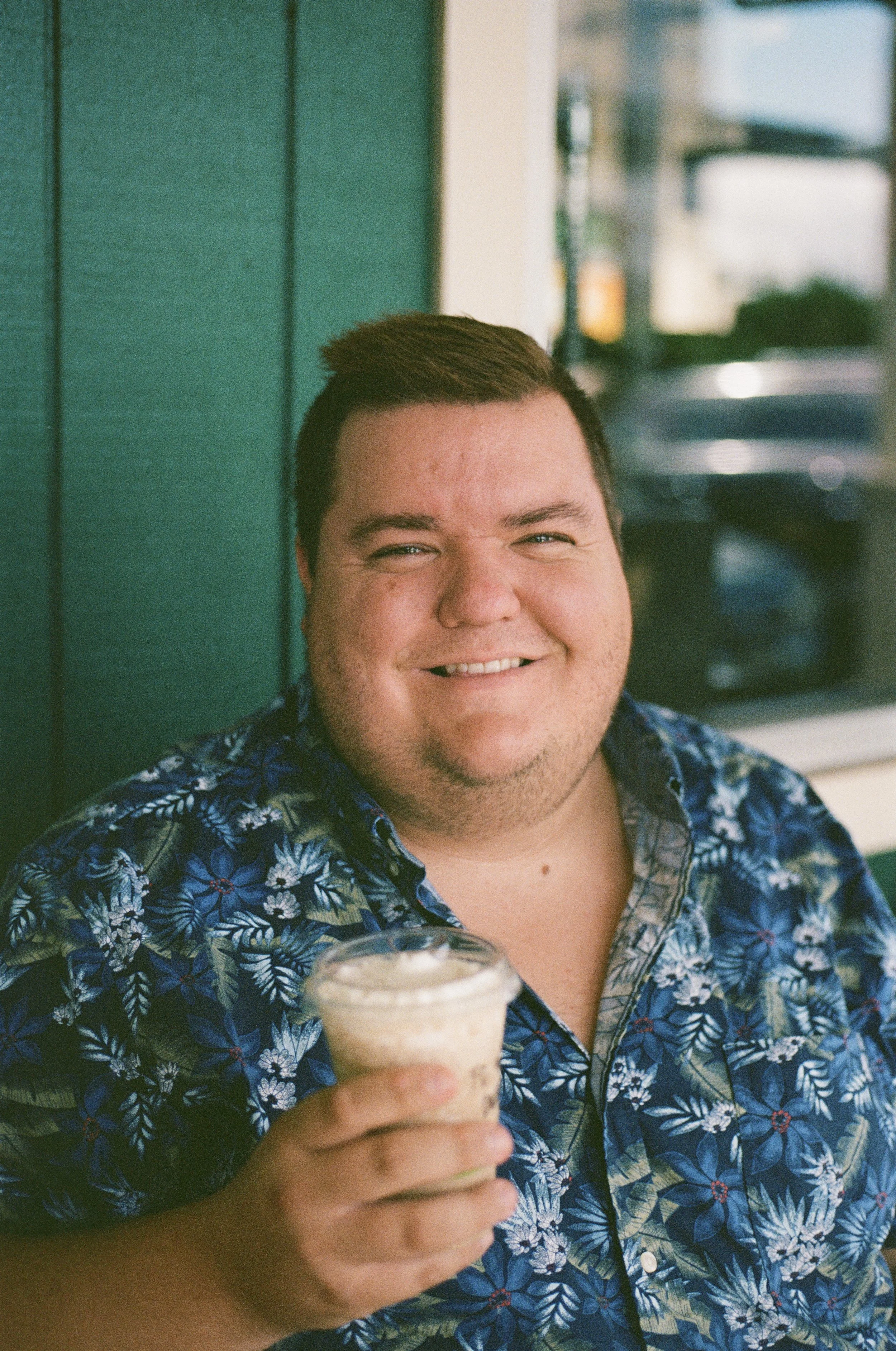 A man smiling and holding a dessert in a cup, sitting outdoors near a green wall.