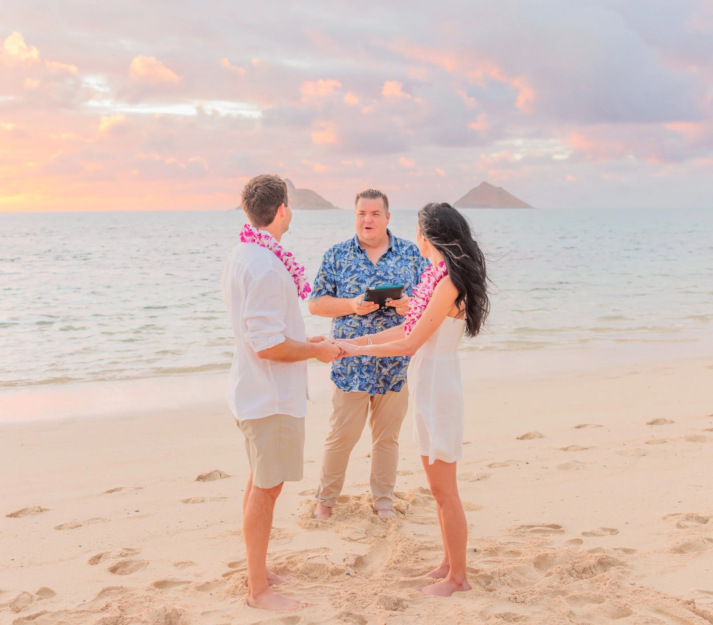 A couple standing on the beach with a woman. They are holding hands and facing each other. A man is holding a small device and seems to be officiating a wedding. The sky is pink and orange with clouds, and there are islands or mountains in the background.