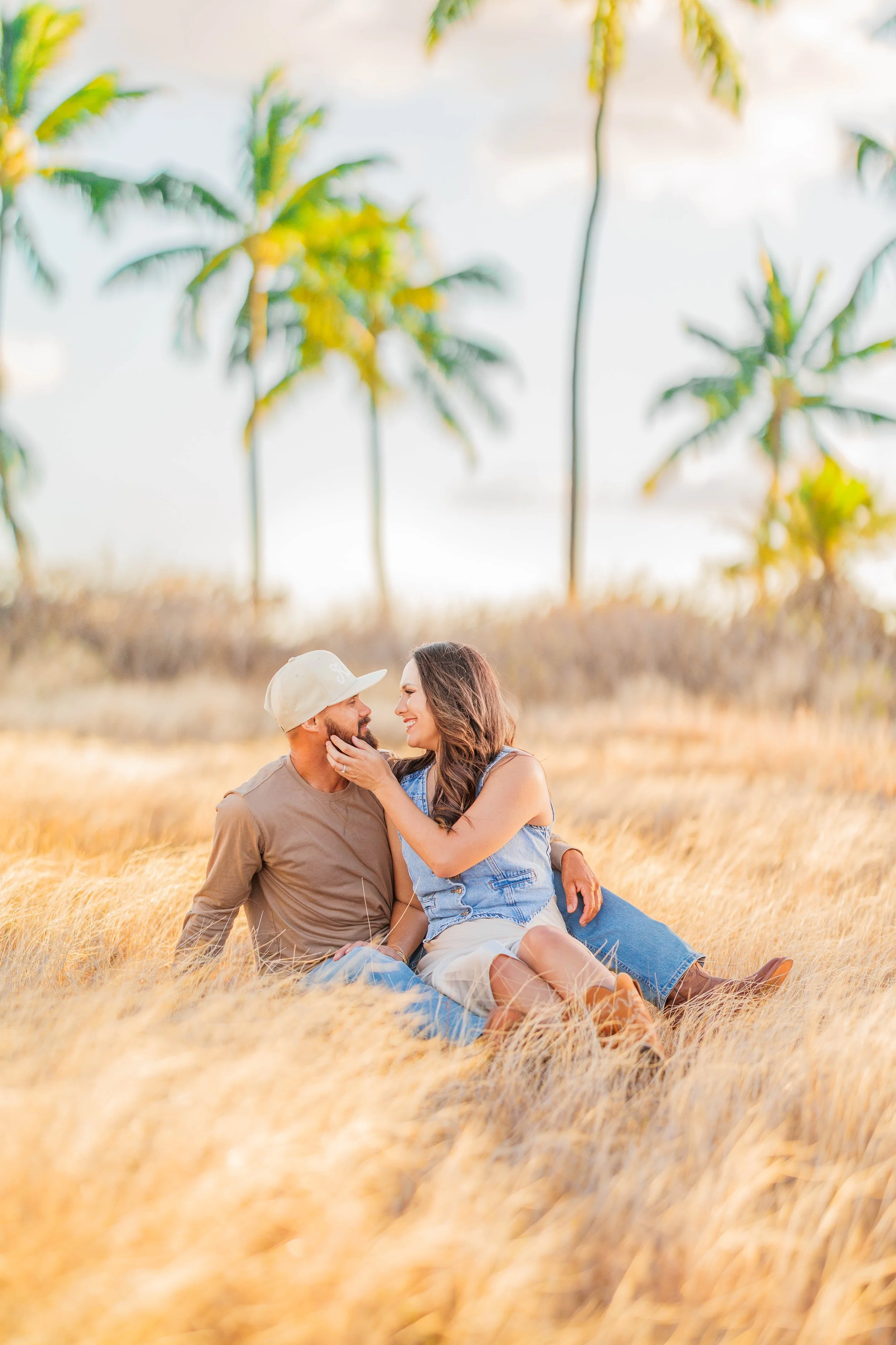 A couple sitting on the grass in a field of tall yellow grass, smiling and looking into each other's eyes, with palm trees in the background during a sunny day.