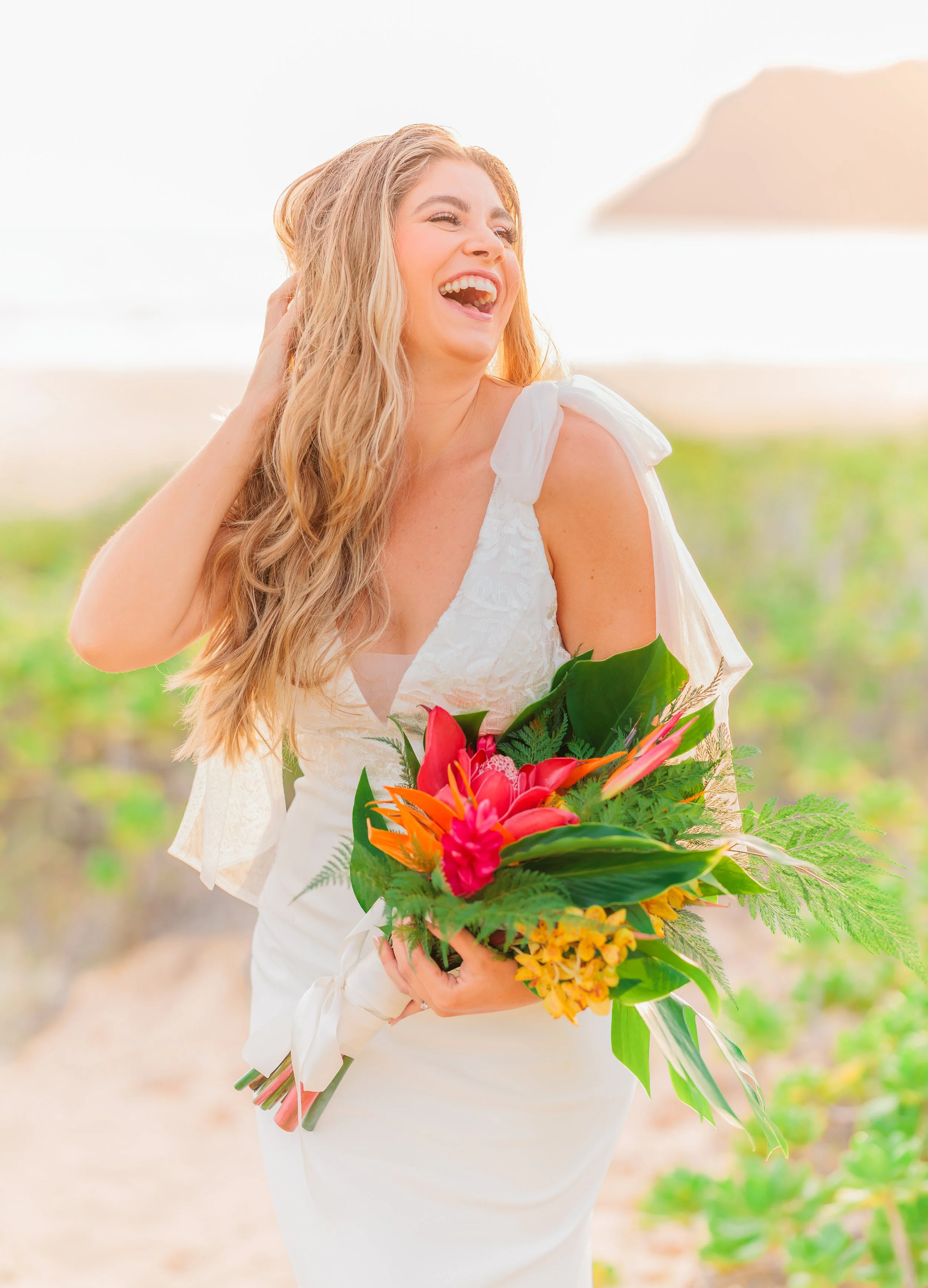 A woman in a white wedding dress holding a colorful tropical bouquet, smiling and laughing outdoors on a sunny day with a beach and green plants in the background.