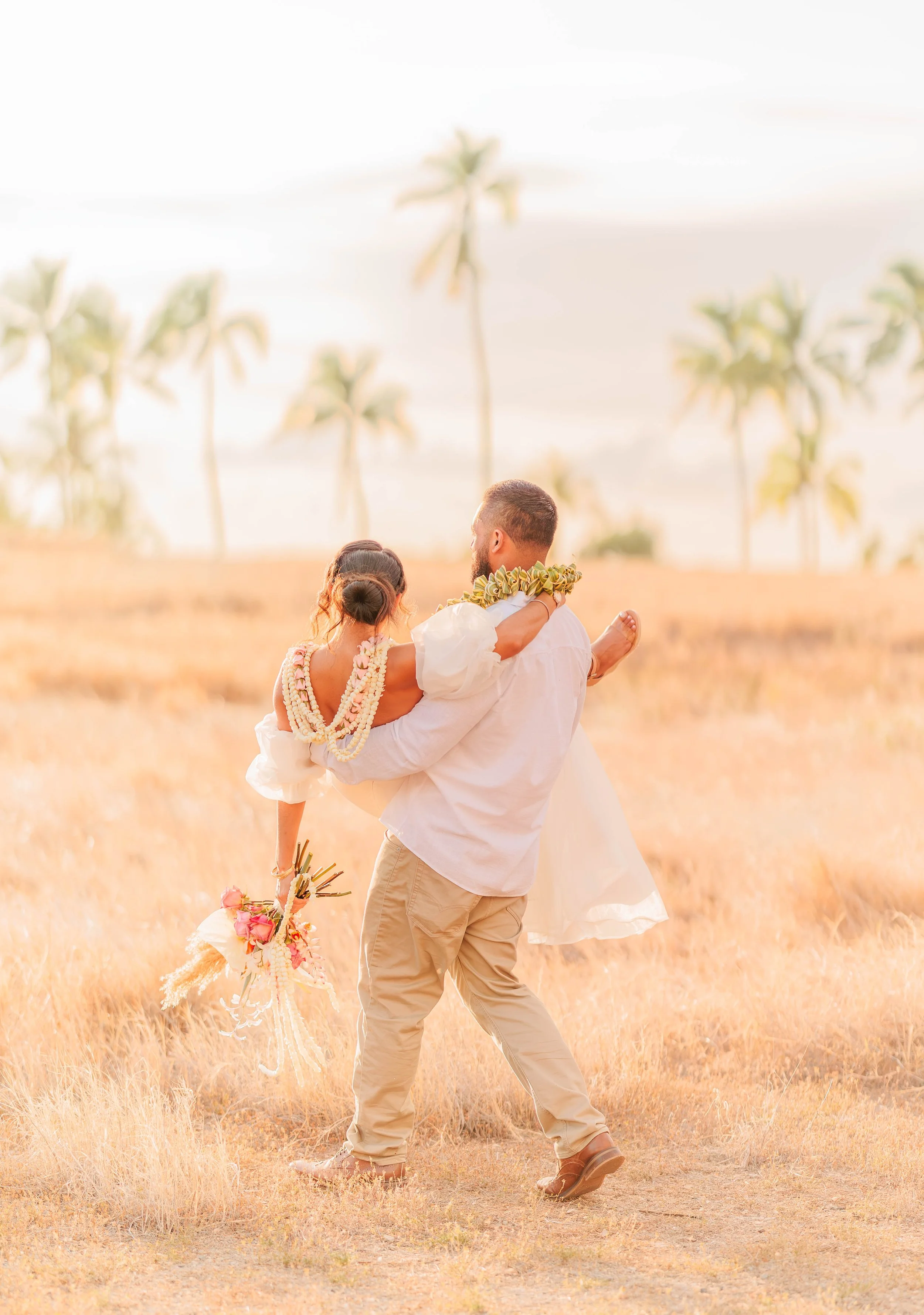 A man holding a woman in a white dress in a grassy outdoor area with palm trees in the background.