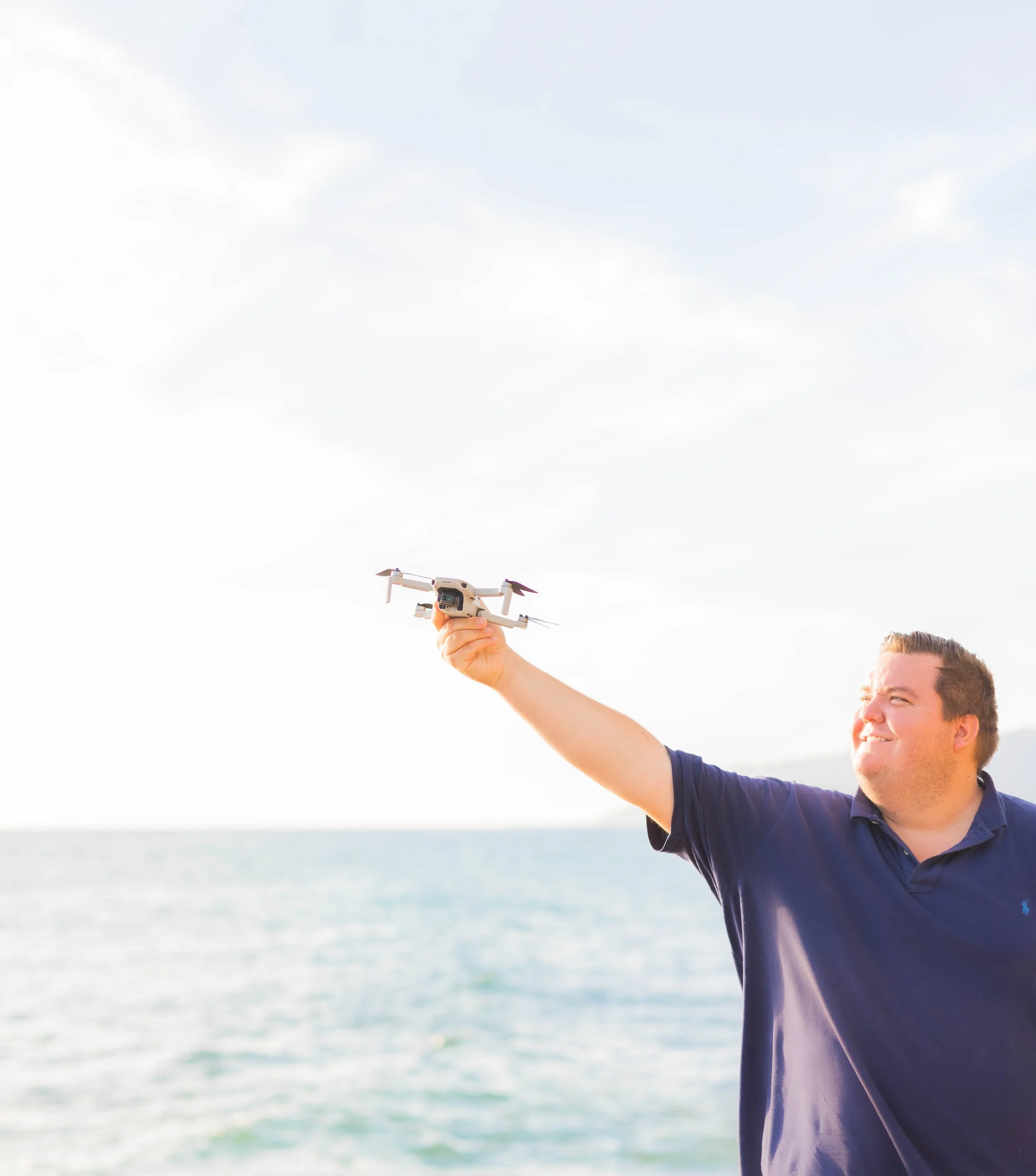 A man in a navy polo shirt holding a drone with the ocean in the background and an overcast sky.