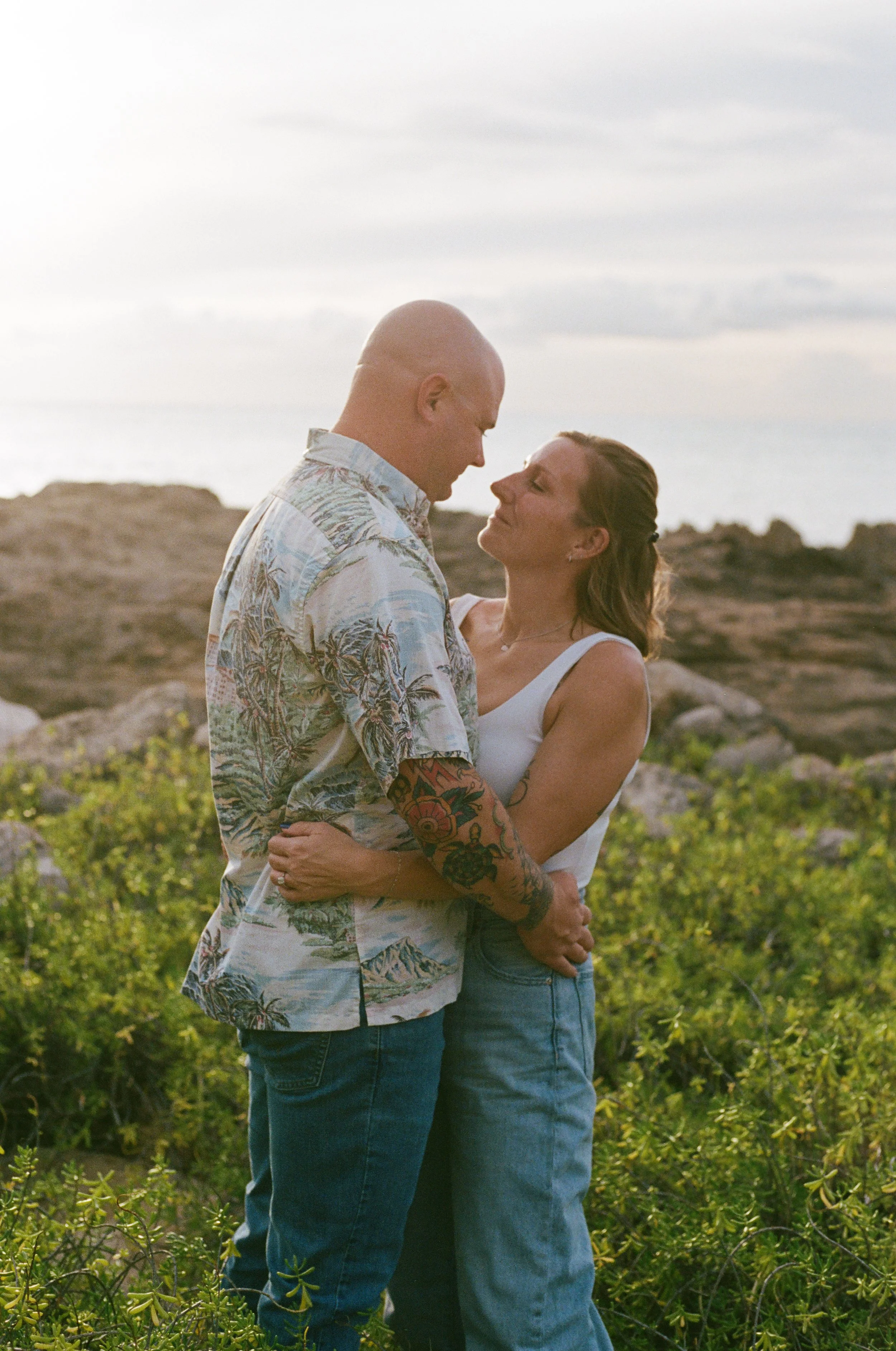 A couple embracing outdoors near the coast, with rocky terrain and shrubs in the background, during sunset.
