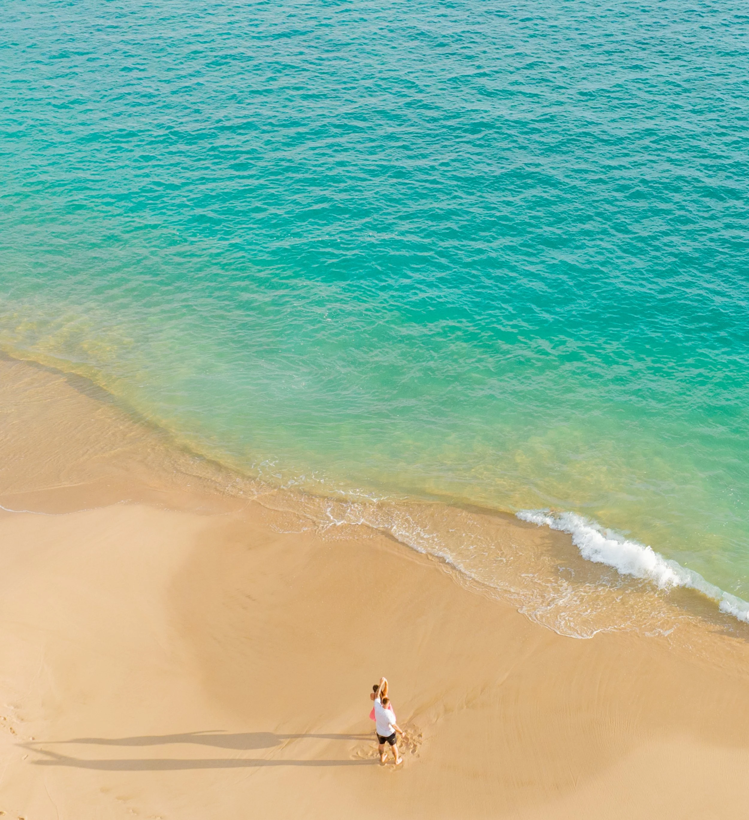 A person walking along a sandy beach with clear blue-green water and waves gently lapping at the shore.