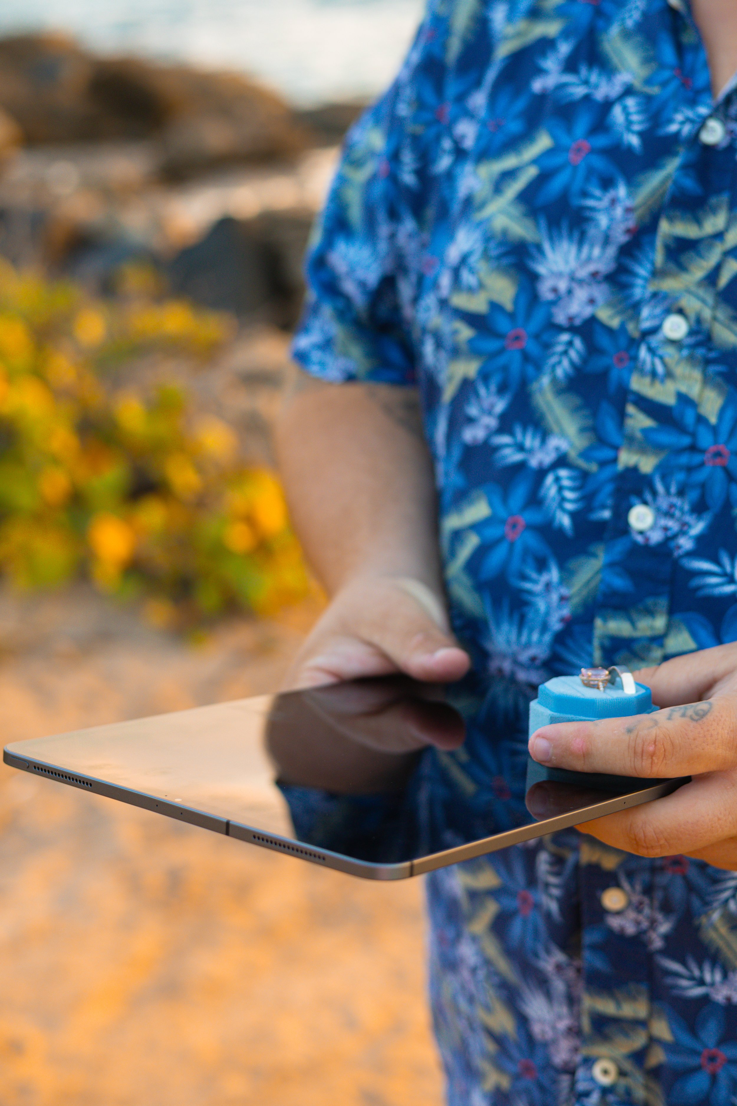 A person in a blue floral shirt holds a tablet while inserting a ring with a small stone into a blue ring box.