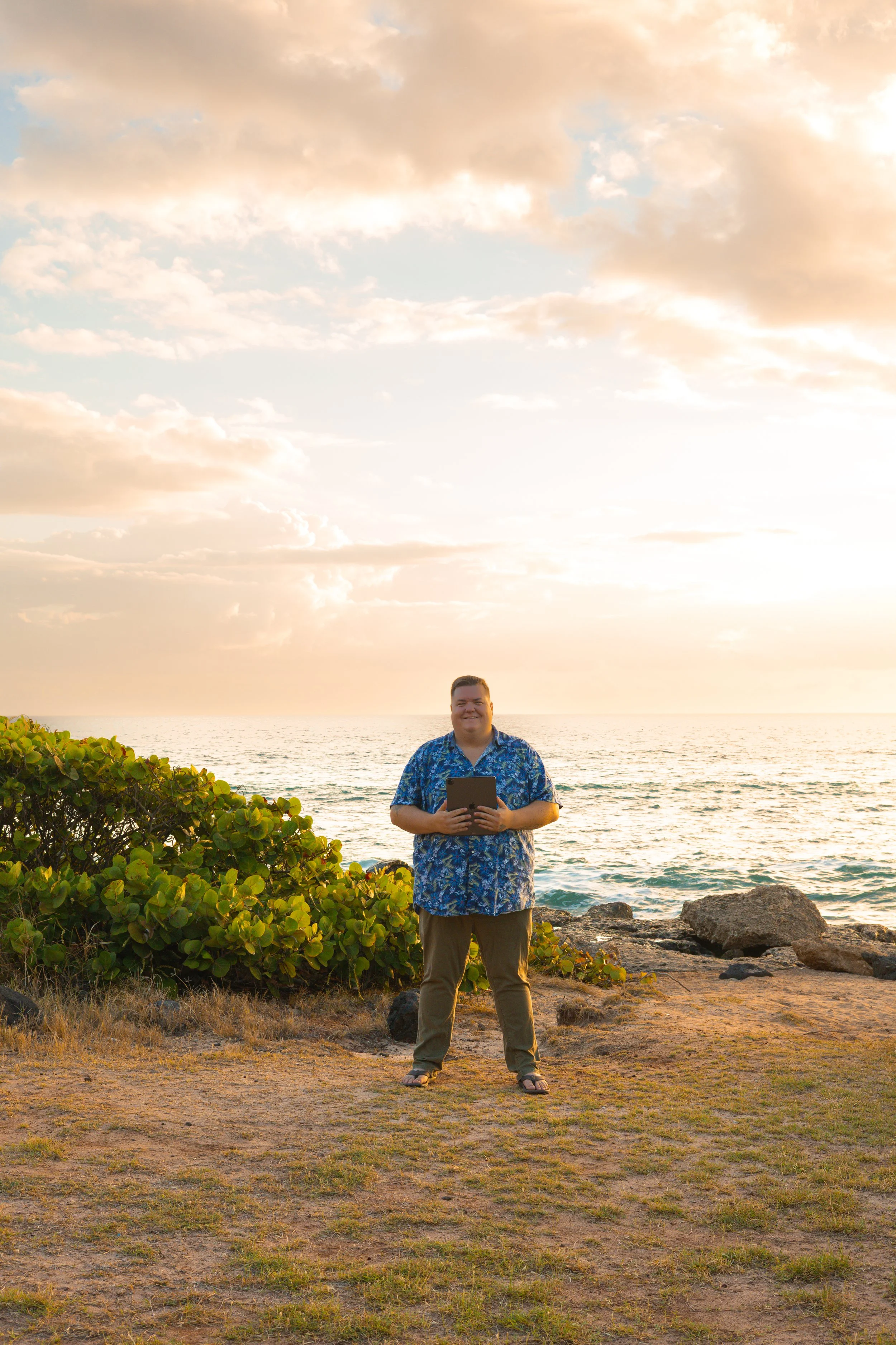 A man standing on the rocky shore at sunset, holding a tablet, with the ocean, rocks, and clouds in the background.