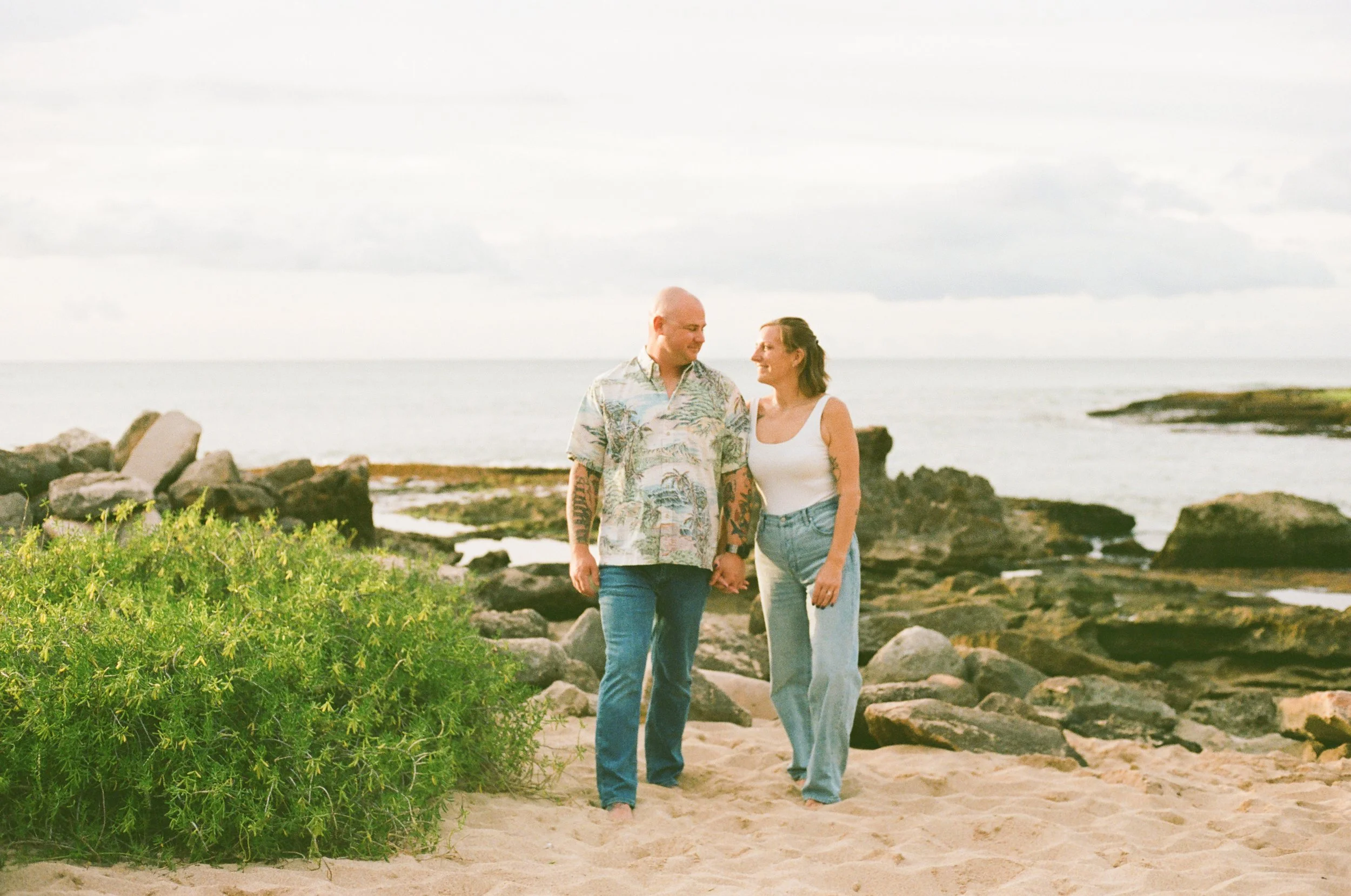 A couple walking hand-in-hand on a sandy beach with rocks and ocean in the background, cloudy sky.