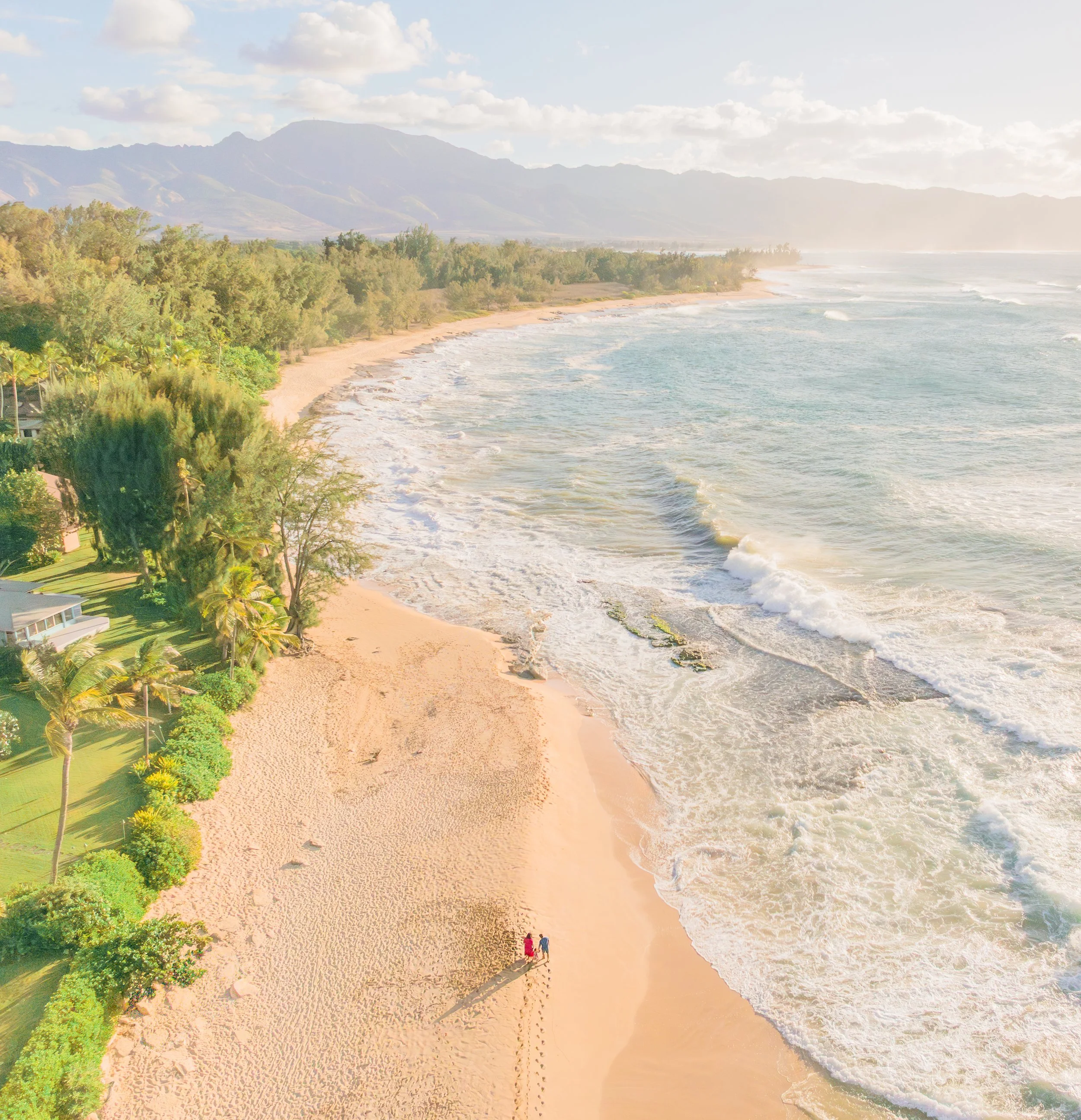 Aerial view of a sandy beach with waves hitting the shore, lined with lush green trees and tropical vegetation, with mountains in the background and a few people walking on the beach.