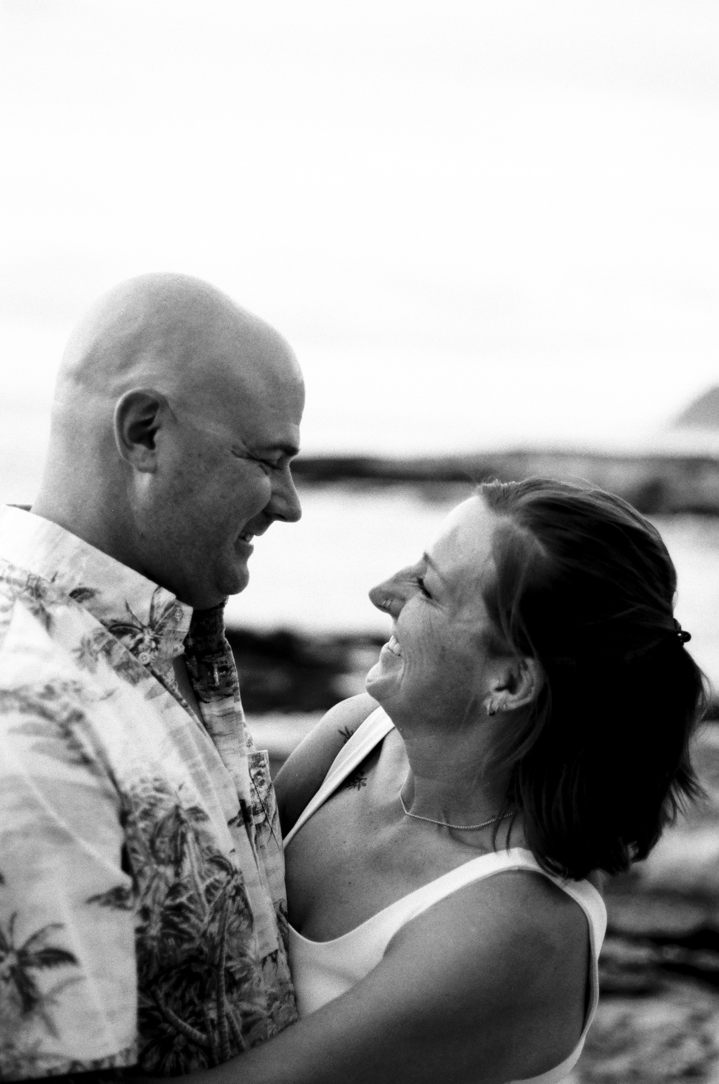 A black-and-white photograph of a smiling couple embracing outdoors, with the sky and distant landscape in the background.
