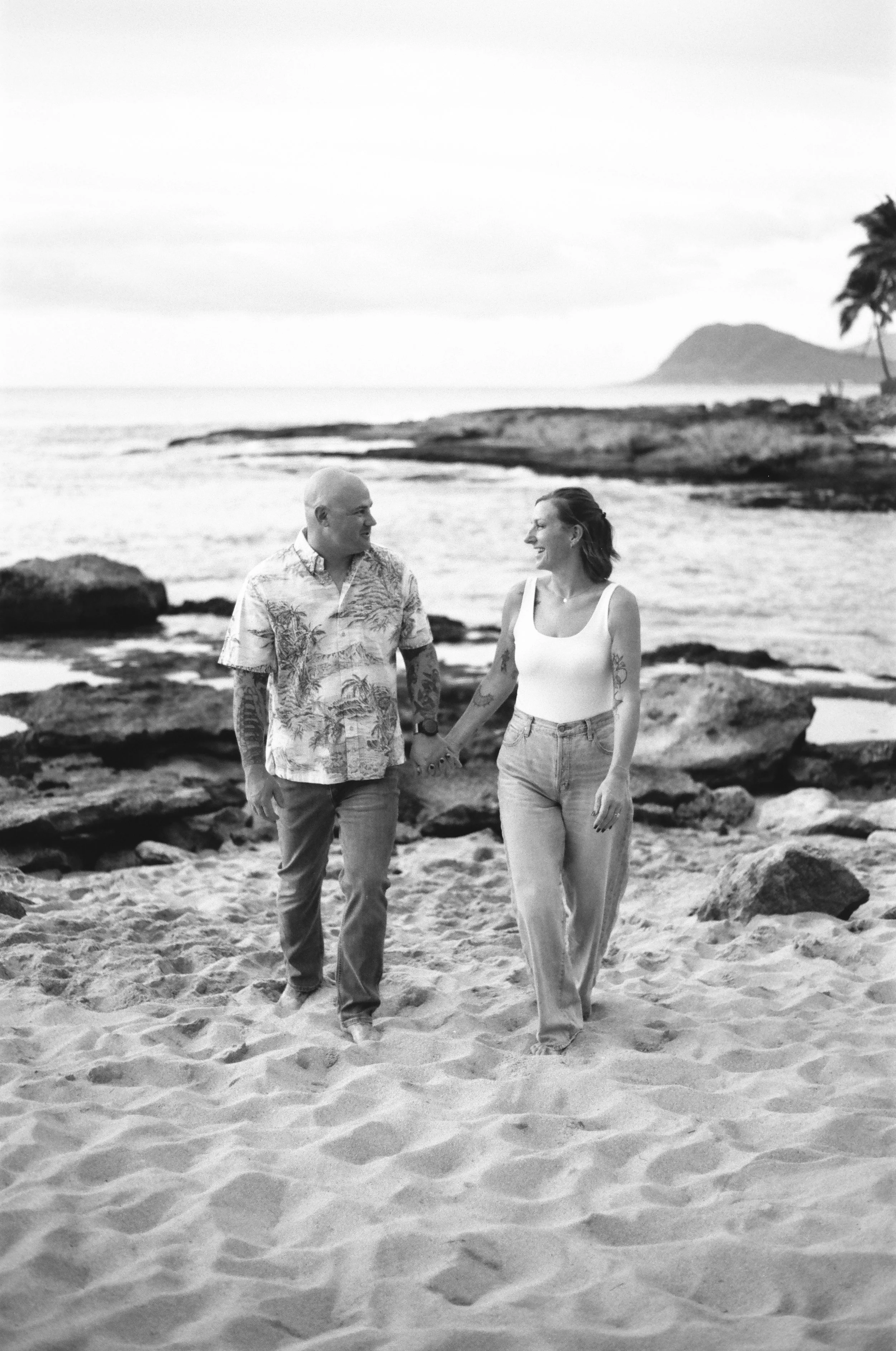 A black-and-white photo of a couple walking on a sandy beach, holding hands and smiling at each other.