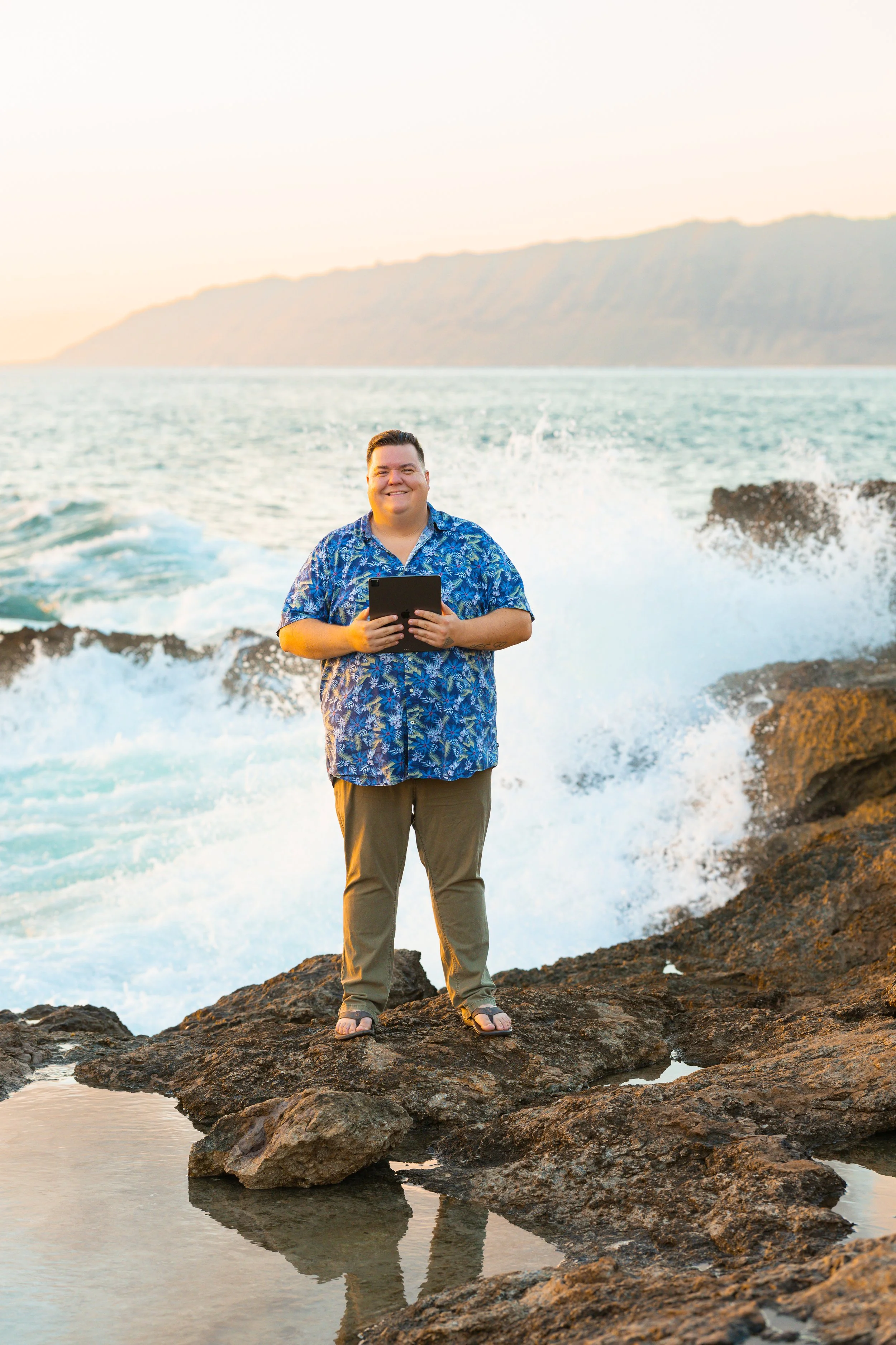 A man in a blue tropical shirt and gray pants standing on rocks by the ocean at sunset, holding a tablet and smiling.