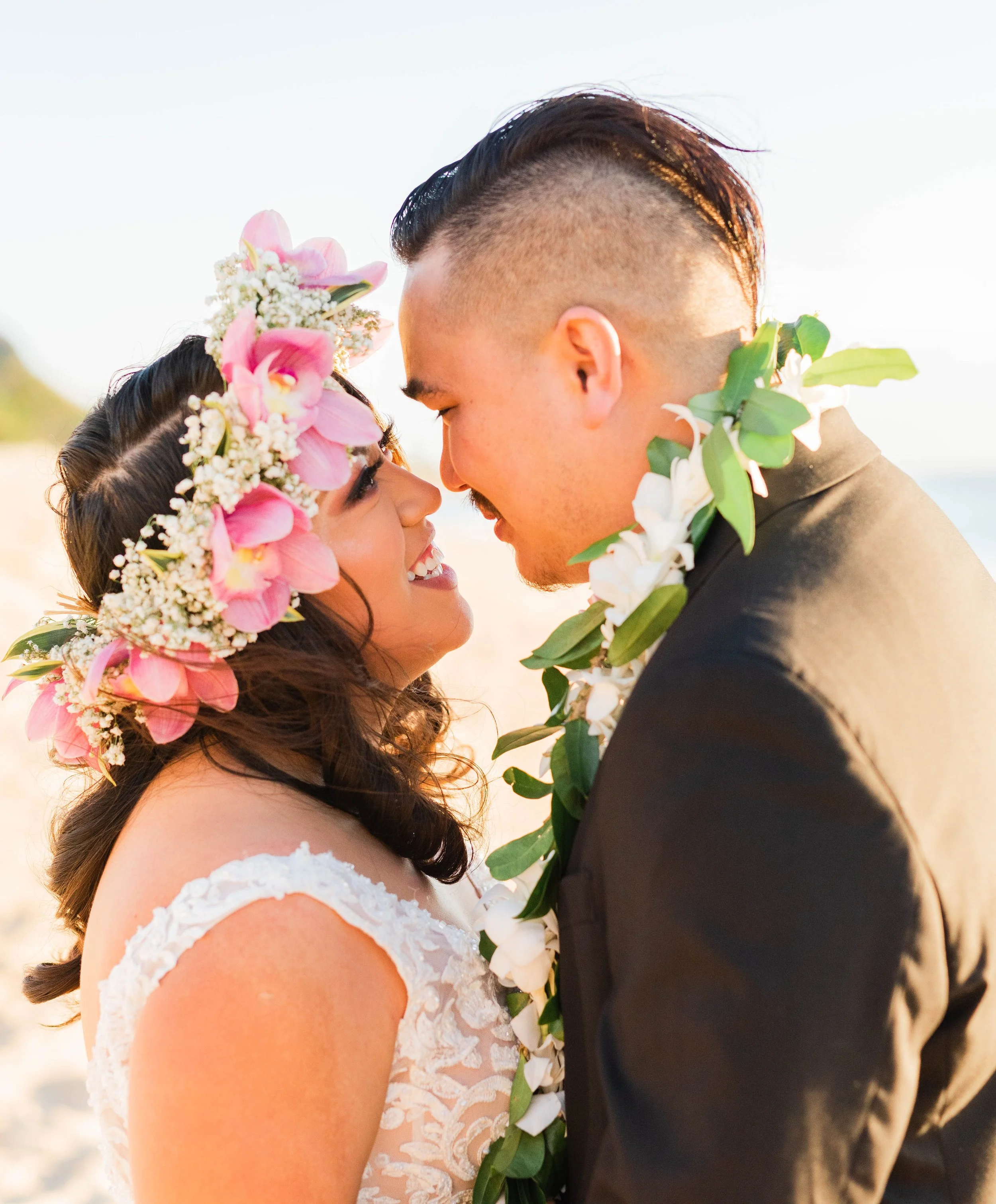 A couple on their wedding day with faces close, smiling, and foreheads touching. The bride is wearing a floral crown, and both are decorated with leis. The background appears to be a beach setting during sunset.
