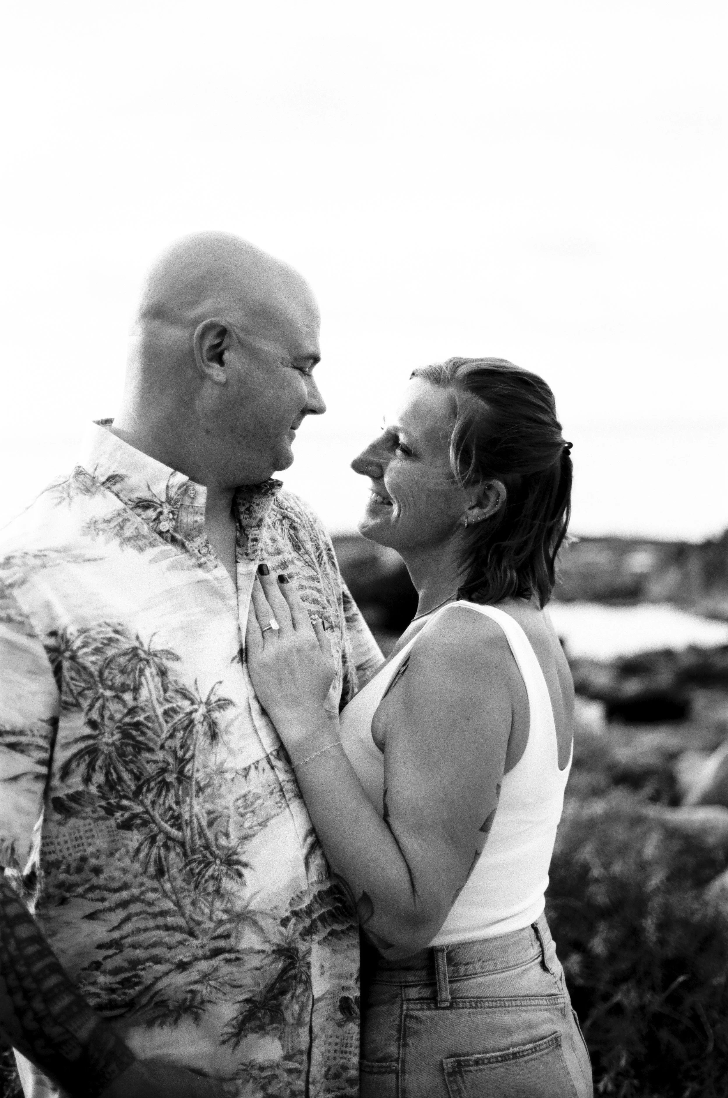 A black-and-white photo of a couple smiling and looking into each other's eyes outdoors.