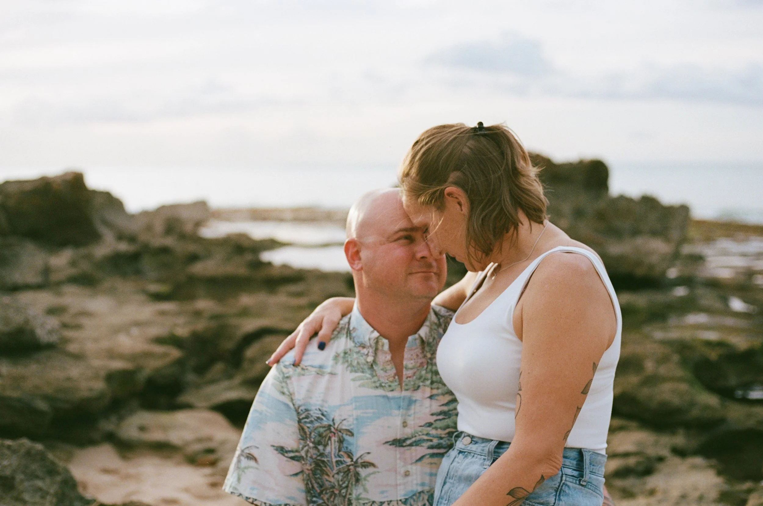 A couple leaning close with foreheads touching on a rocky beach, the man in a colorful short-sleeve shirt and the woman in a white tank top and denim shorts, both with tattoos and embracing each other.