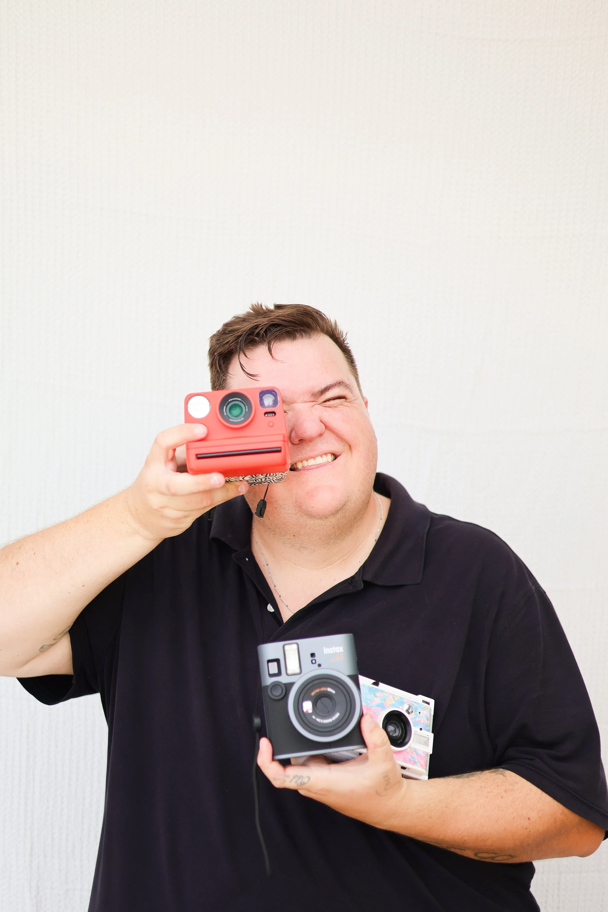 A man smiling and holding up a Polaroid camera, with a box of instant film and a small camera in his hands. He is wearing a black polo shirt and is standing against a plain white wall.