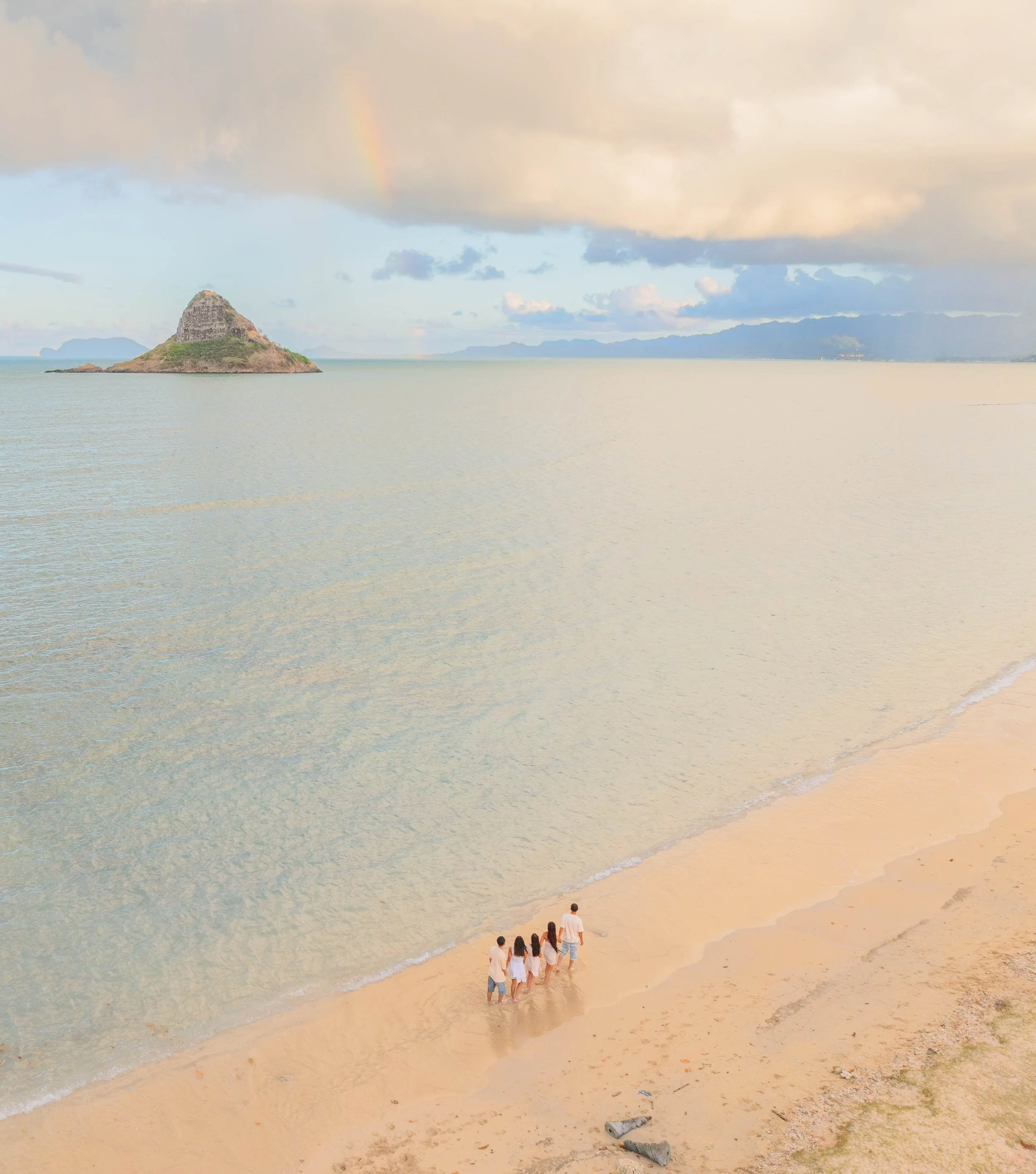 A family walking along the beach at sunset with a small island and rainbow in the background.