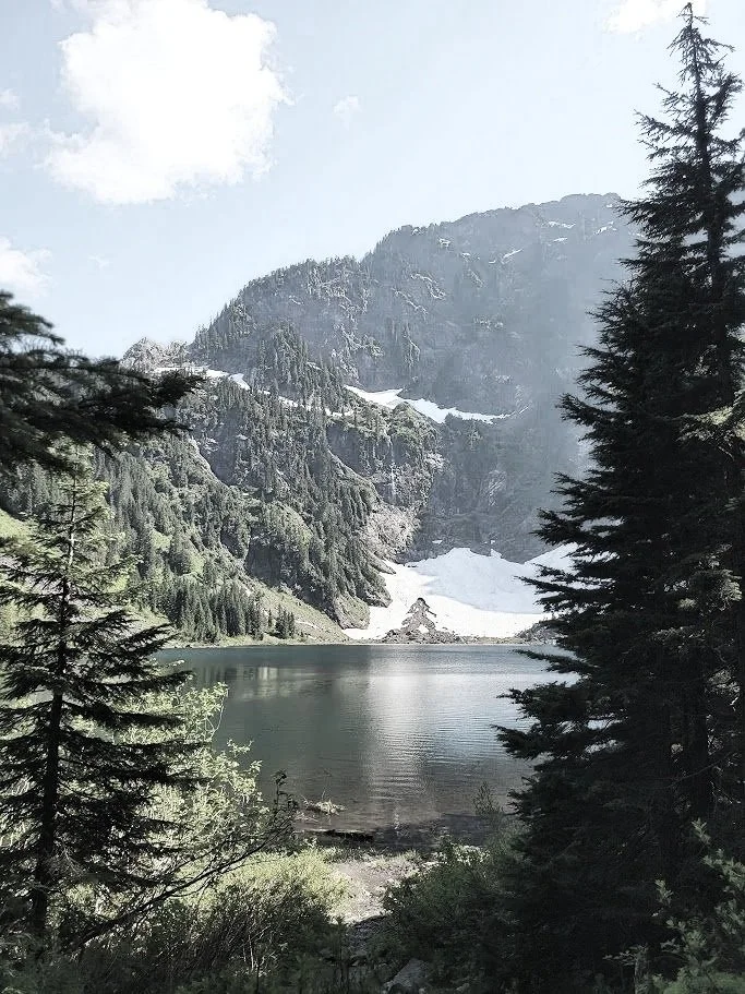 Mountain lake surrounded by evergreen trees with snow patches on the mountain slopes under a partly cloudy sky.