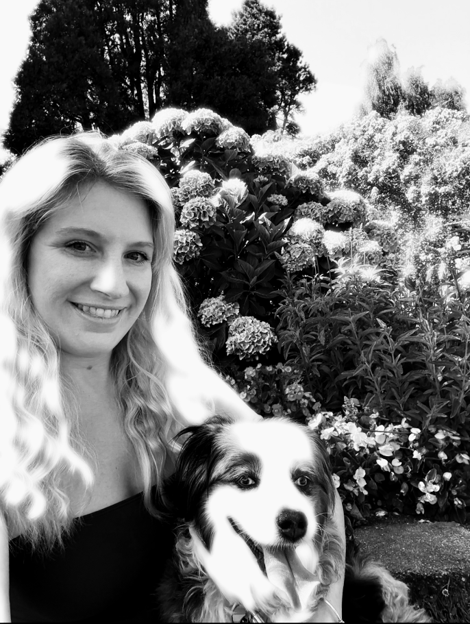 A woman with long wavy hair smiling next to a happy dog with its tongue out, surrounded by blooming flowers and trees.