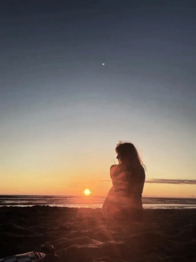 A person sitting on the beach at sunset, facing the ocean with the sun low on the horizon, and a star visible in the sky.
